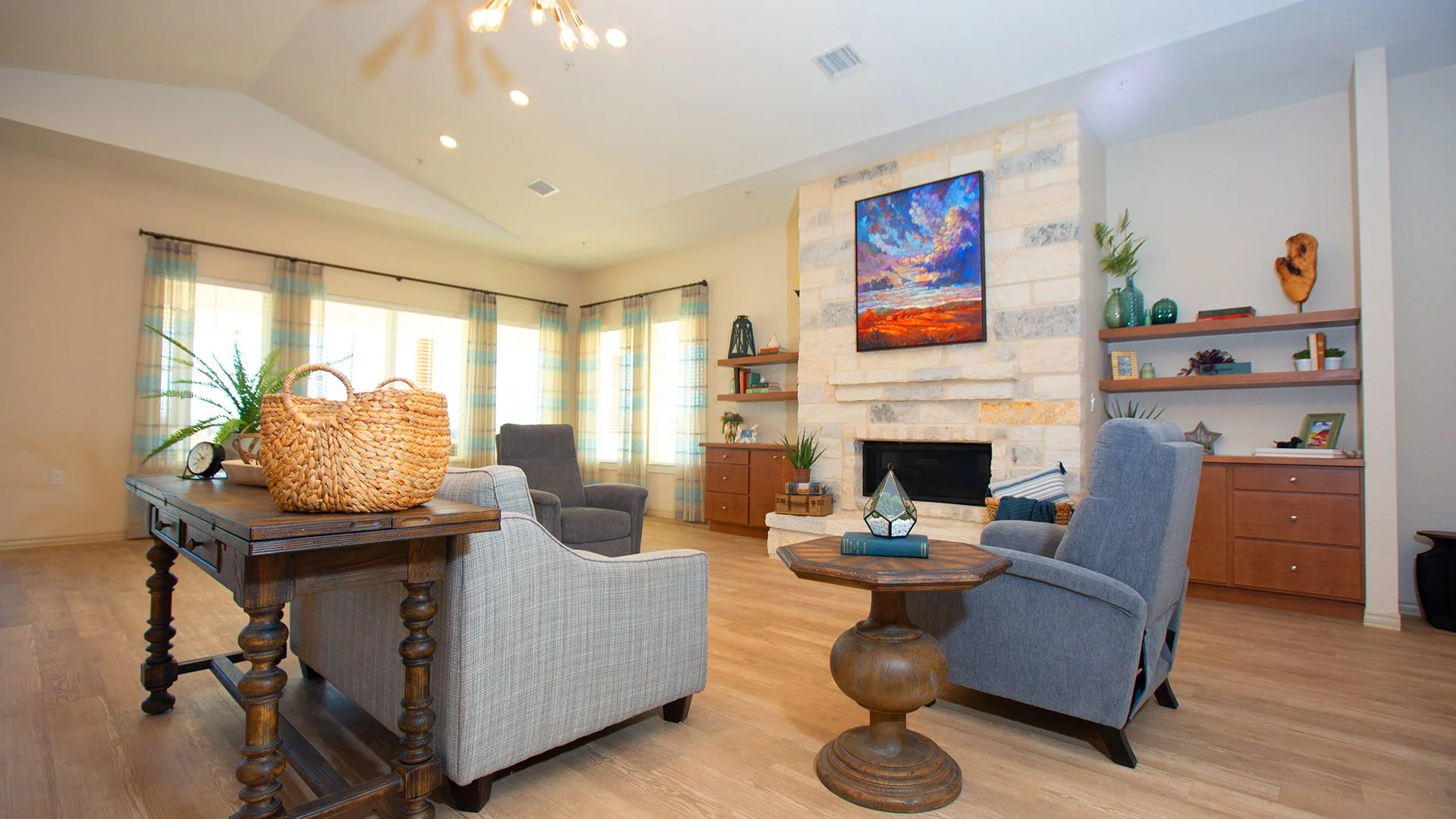Living room with a stone fireplace, colorful painting above it, wooden and upholstered furniture, decorative plants, and a large window with striped curtains.