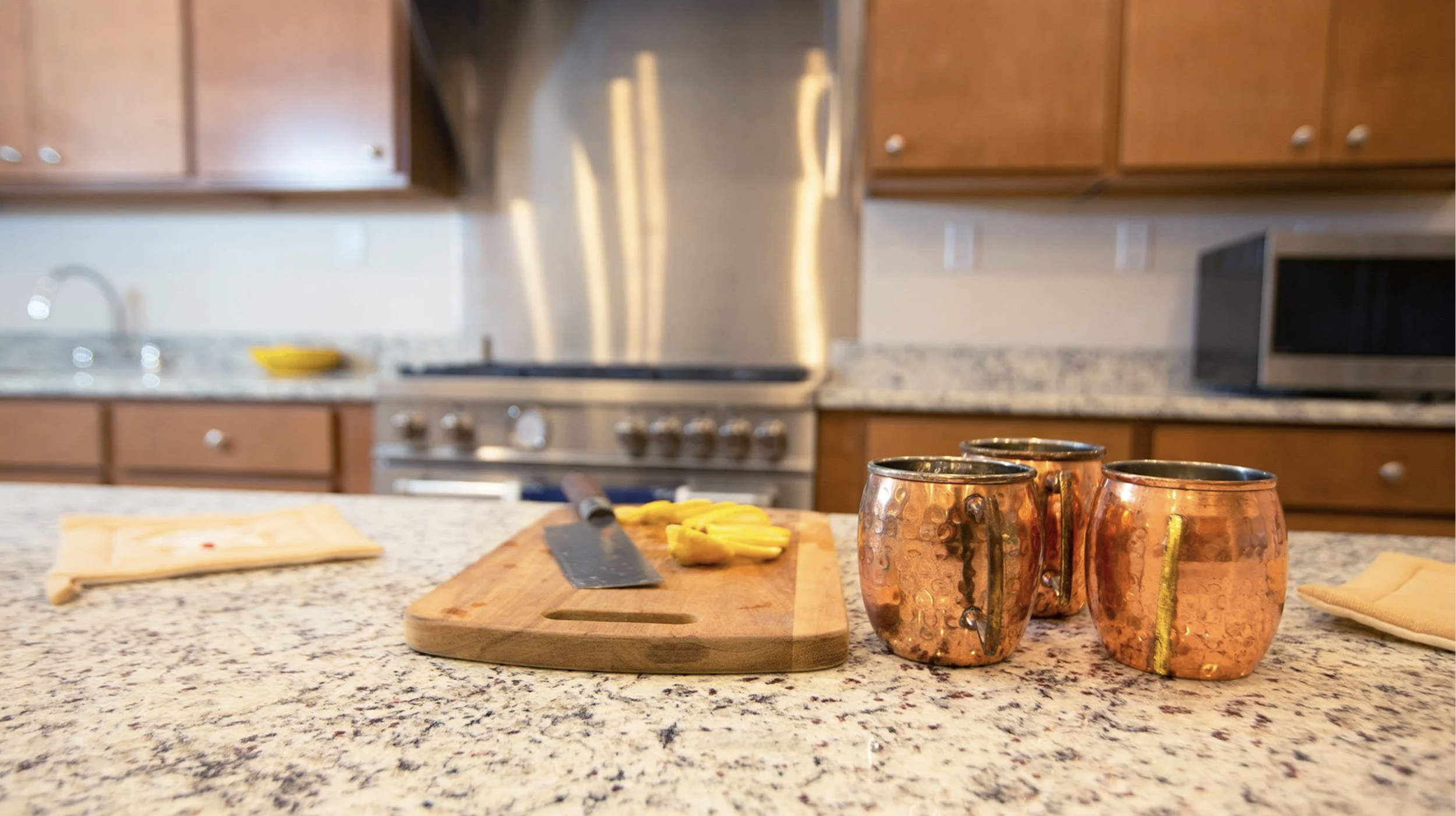 Kitchen countertop with three copper mugs, a wooden cutting board with sliced yellow peppers, a knife, a folded cloth, and a napkin in the foreground; kitchen cabinets, stove, microwave, and a silver backsplash in the background.