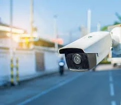 A security camera mounted outdoors on a pole brewing over a street with a cloudy sky.