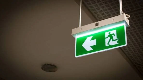 Green emergency exit sign with a running figure and an arrow pointing to the left, hanging from the ceiling.