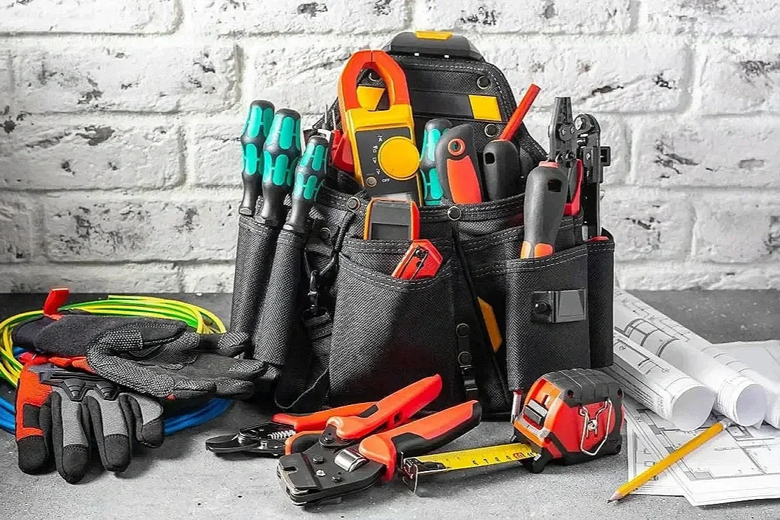 Tool belt filled with various hand tools, measuring tapes, gloves, and electrical testers on a work surface with rolled blueprints and pencils.