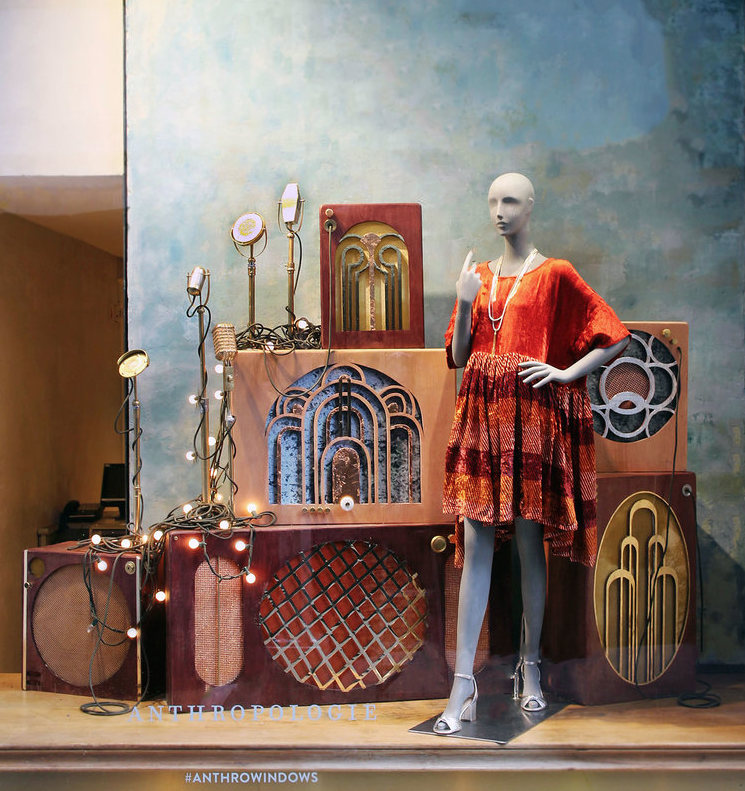 A mannequin dressed in an orange patterned dress with jewelry, standing in front of a display of vintage-style radio speakers and lights in a store or showroom. Retail window display by Breena Farrelly for Anthropologie.