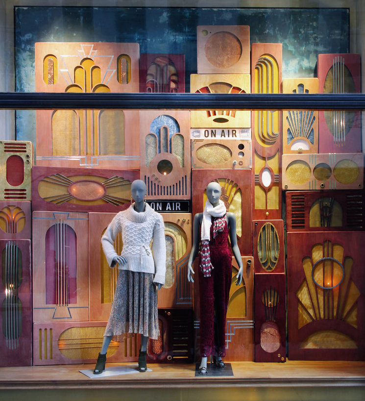 Display window with two mannequins dressed in winter clothing, set against a colorful backdrop of abstract geometric shapes resembling vintage radios with 'ON AIR' signs. Retail window display by Breena Farrelly for Anthropologie.