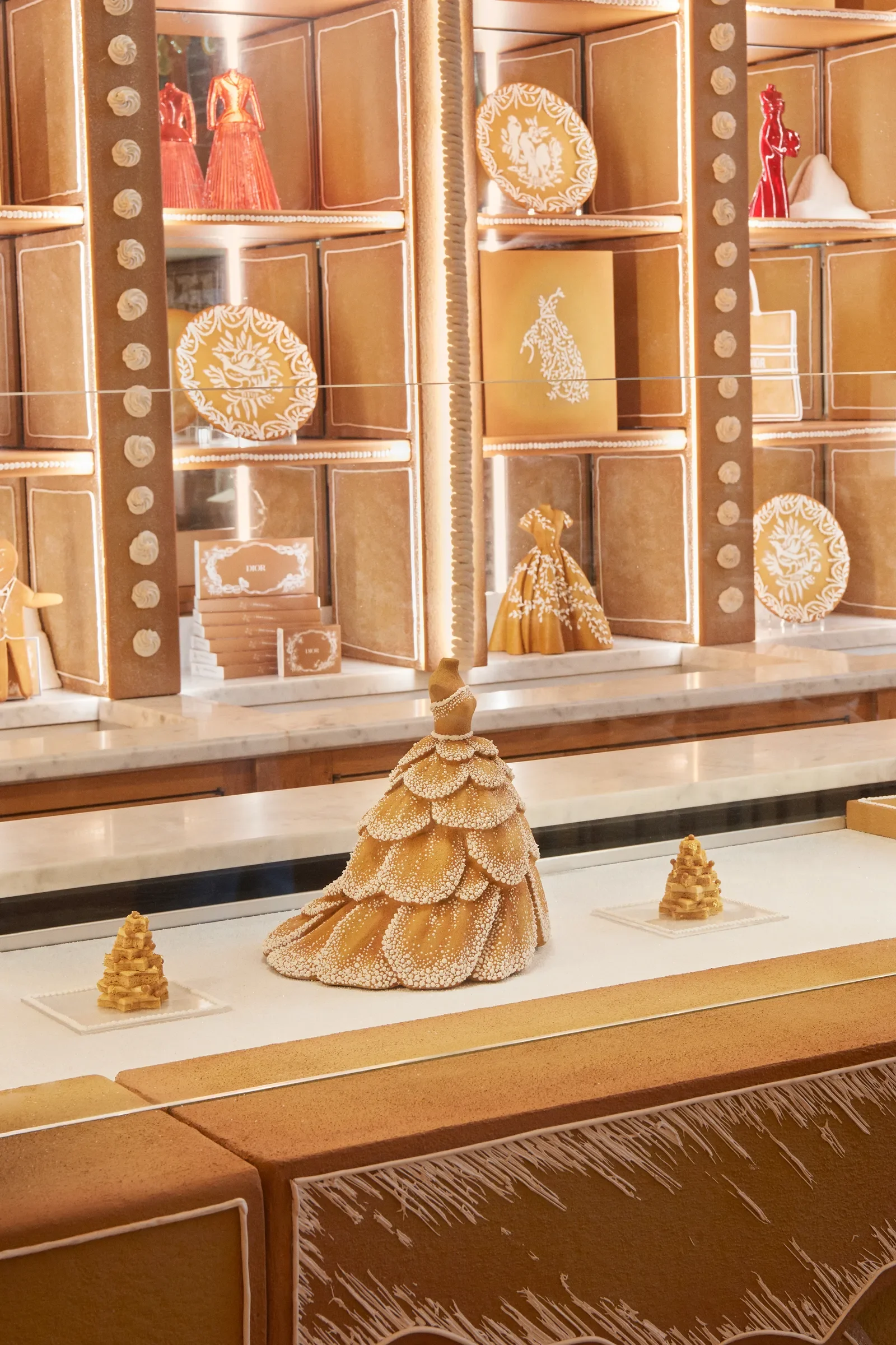 Decorative gingerbread display featuring a large gingerbread tree centerpiece and two smaller gingerbread trees, with a backdrop of gold and white Christmas-themed ornaments and packaging.