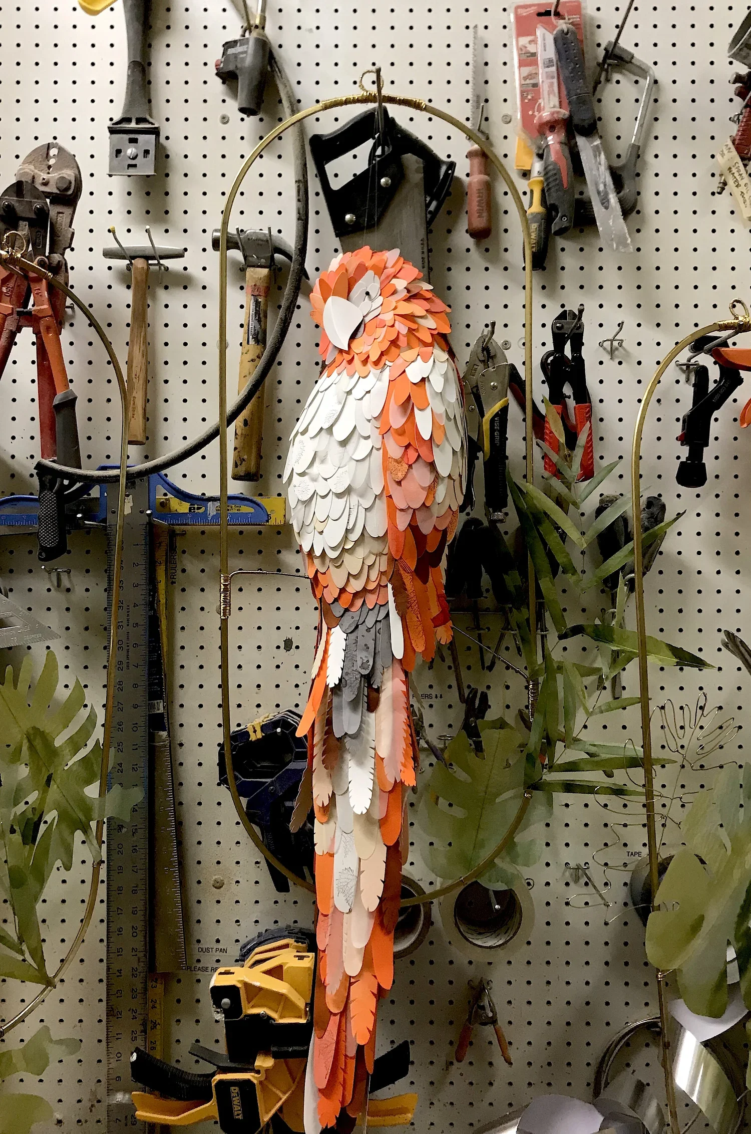 A decorative parrot made of layered orange, white, gray, and beige paper or fabric feathers hanging on a metal stand in a workshop with various tools and green plant leaves in the background.