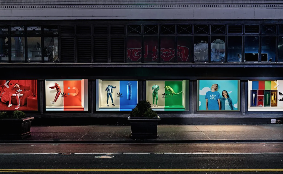 Display window with colorful athletic apparel and accessories, including mannequins dressed in Adidas clothing, some with sports equipment, and a backdrop with models wearing Adidas in natural settings, on a city street at night.
