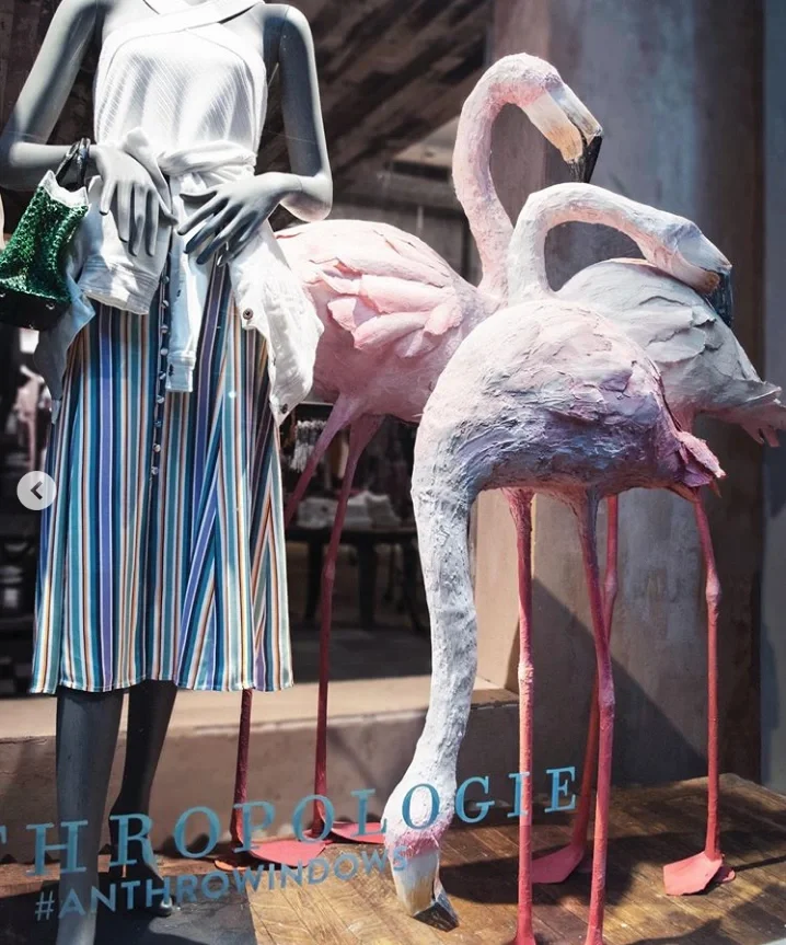 Display of a mannequin dressed in a white top and multicolored striped skirt, set next to three pink flamingo statues, with the words 'ANTHROPOLOGIE' and '#ANTHROWINDOWS' at the bottom.