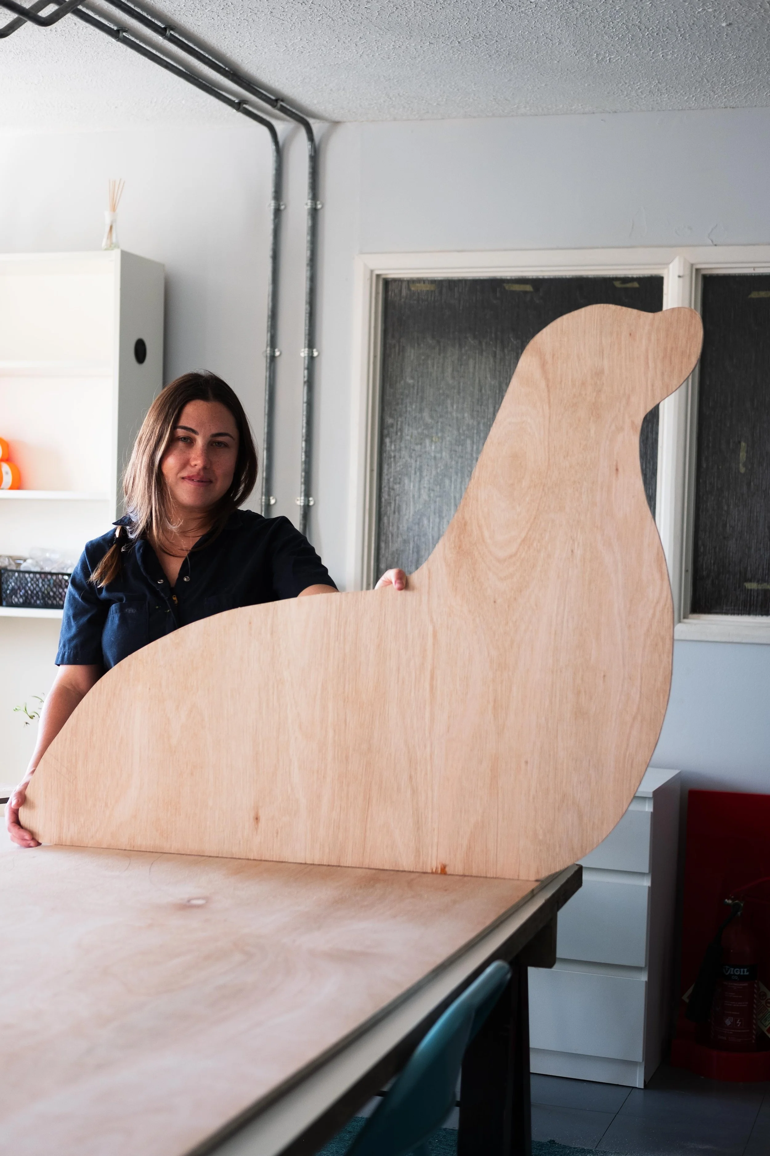 Breena Farrelly Props & Displays holding a large unfinished wooden cutout of a dog while standing at a table in a room with white walls and a window.