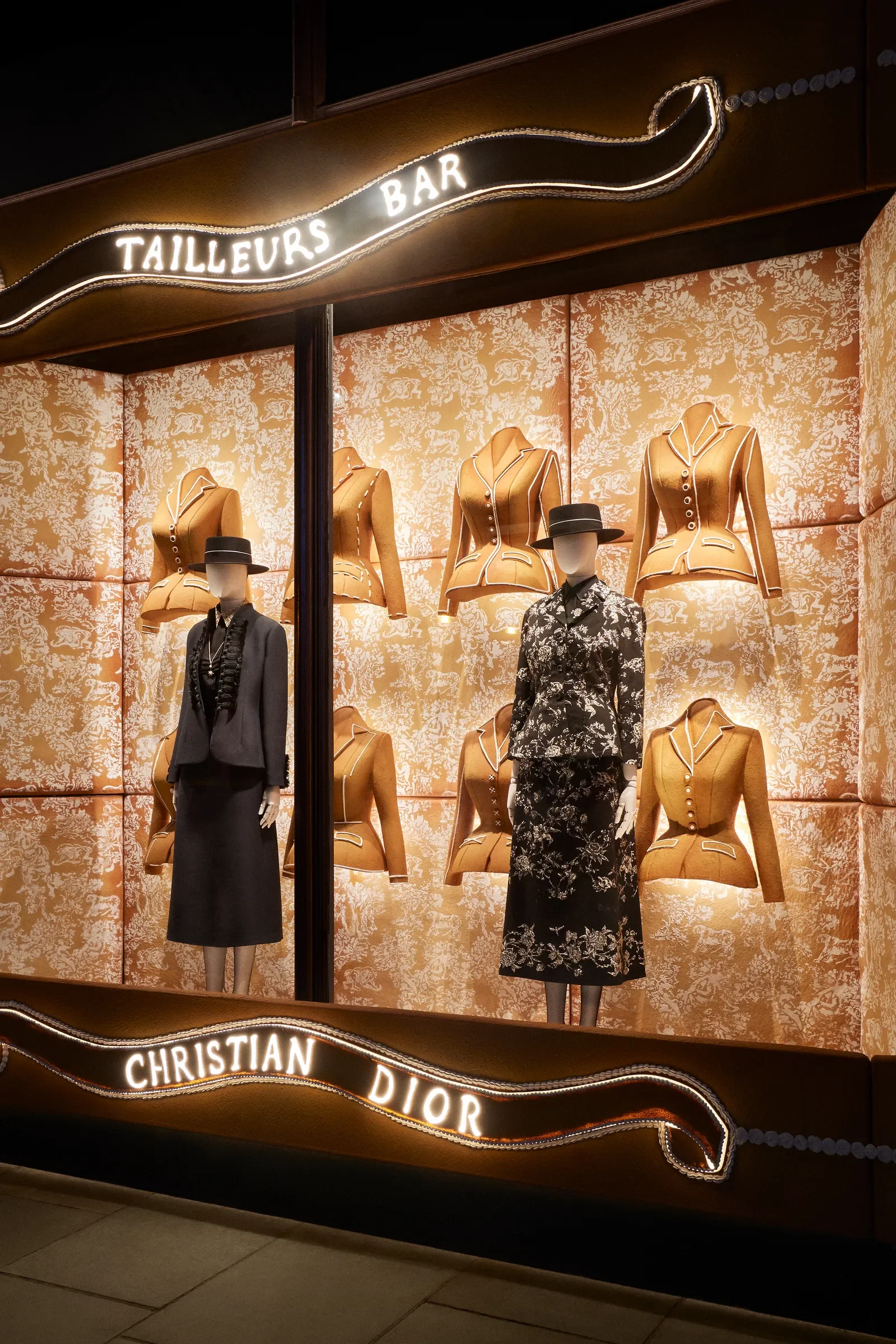 Display window for Christian Dior featuring mannequins dressed in vintage-style women's clothing with a background of illuminated tailored jackets. Breena Farrelly Props & Displays.