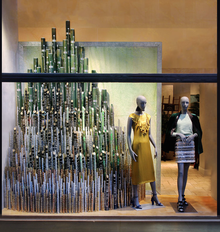 Clothing store display with mannequins wearing yellow and striped outfits, and a decorative structure made of numerous green and gold vertical rods in the background. Retail window display by Breena Farrelly for Anthropologie.