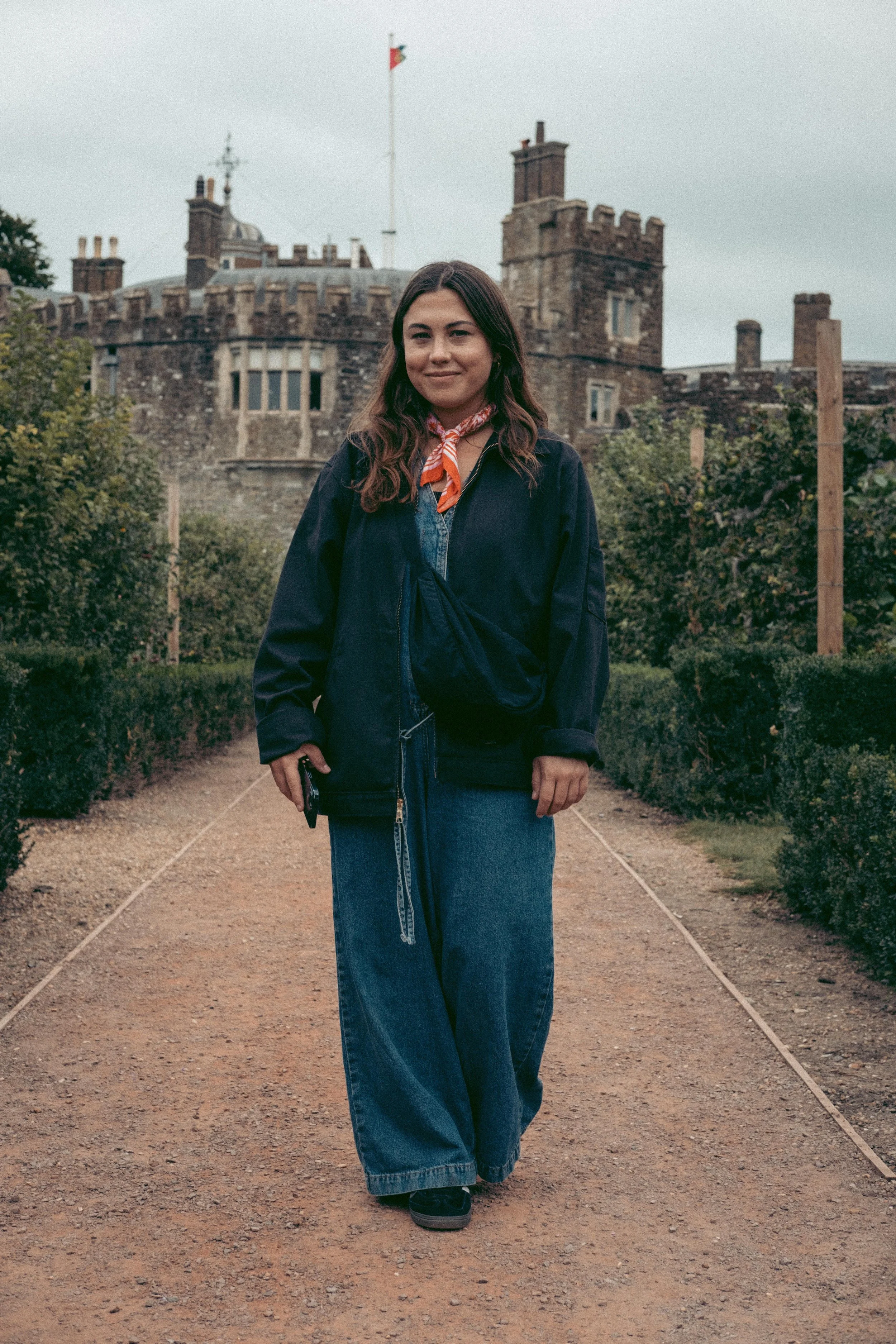 A young woman (Breena Props & Displays) with long wavy hair standing on a gravel path in front of a historic castle (Walmer Castle), wearing a dark jacket, wide jeans, and an orange and white scarf, holding a phone in her right hand.