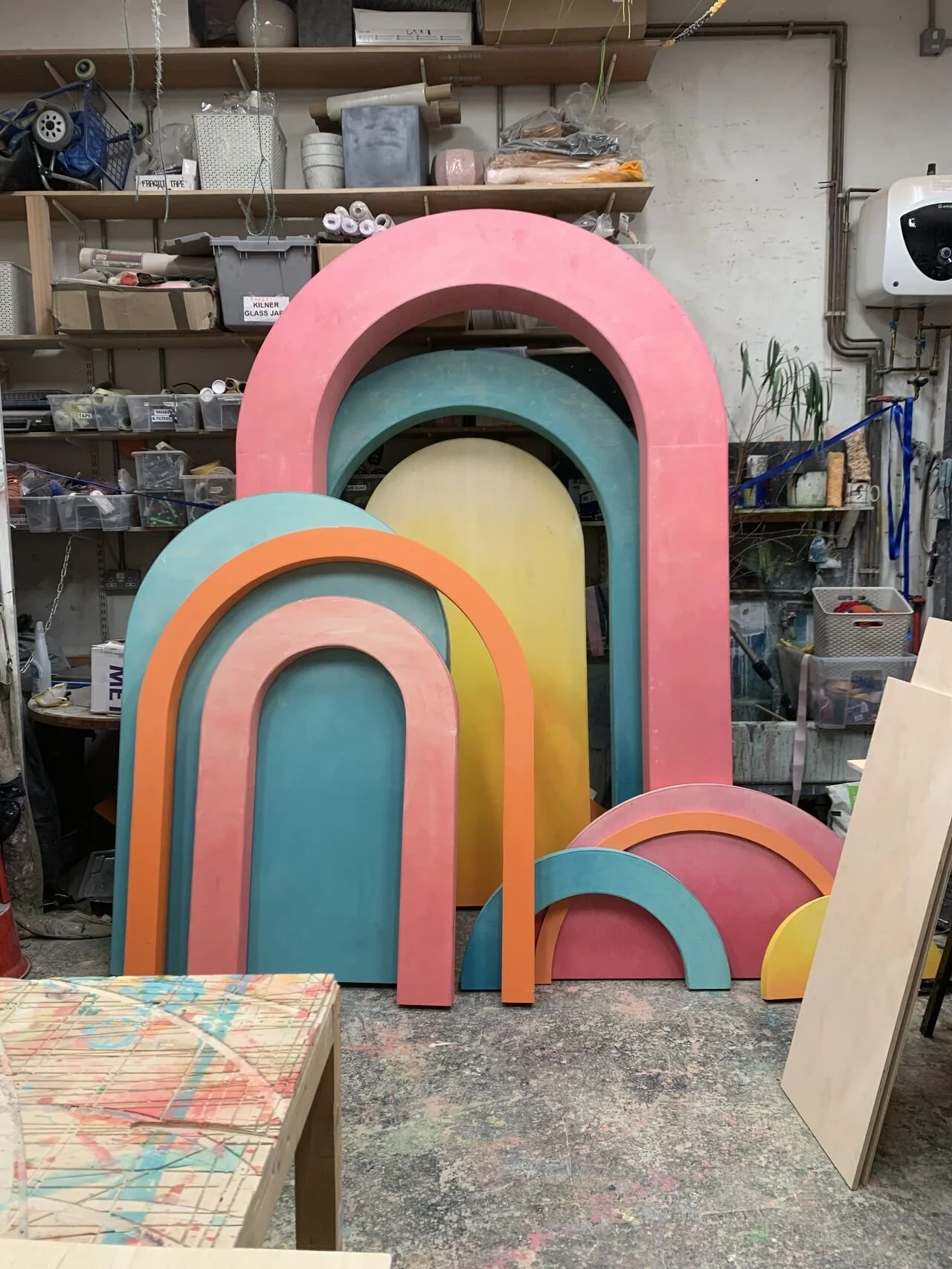 Colorful rainbow-shaped wooden cutouts in pastel colors including pink, teal, yellow, and orange in an artist's workshop or storage space.