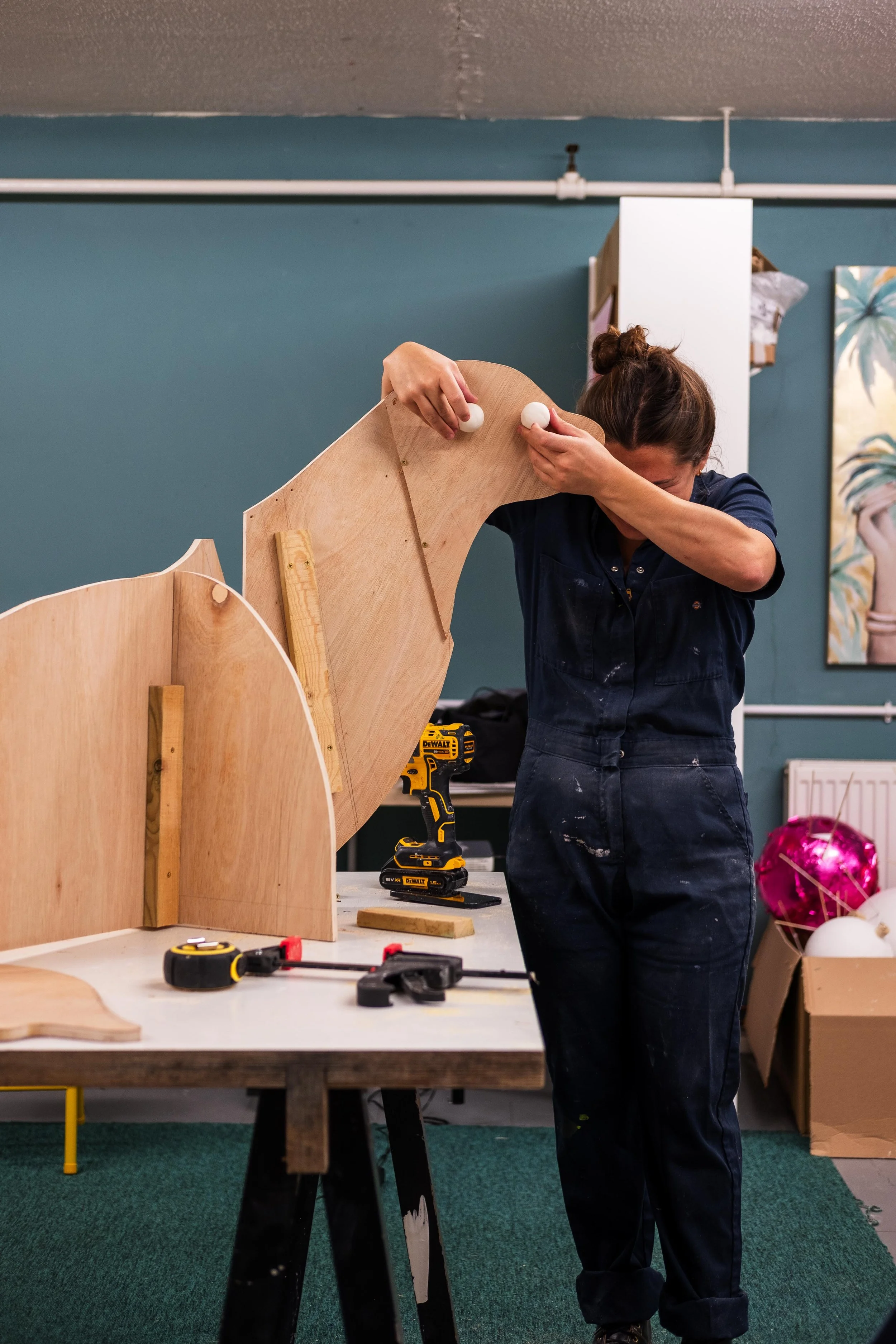Breena Farrelly Props & Displays working on assembling a wooden piece on a worktable using a drill, with other tools and materials on the table, in a workshop setting.