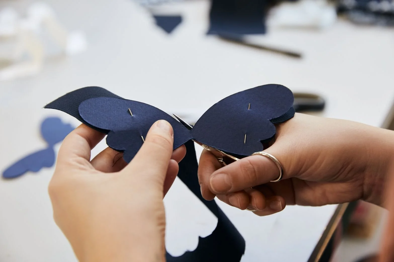 Two hands holding a dark blue paper butterfly with visible pins, amid a workspace with additional paper butterflies and crafting tools in the background.