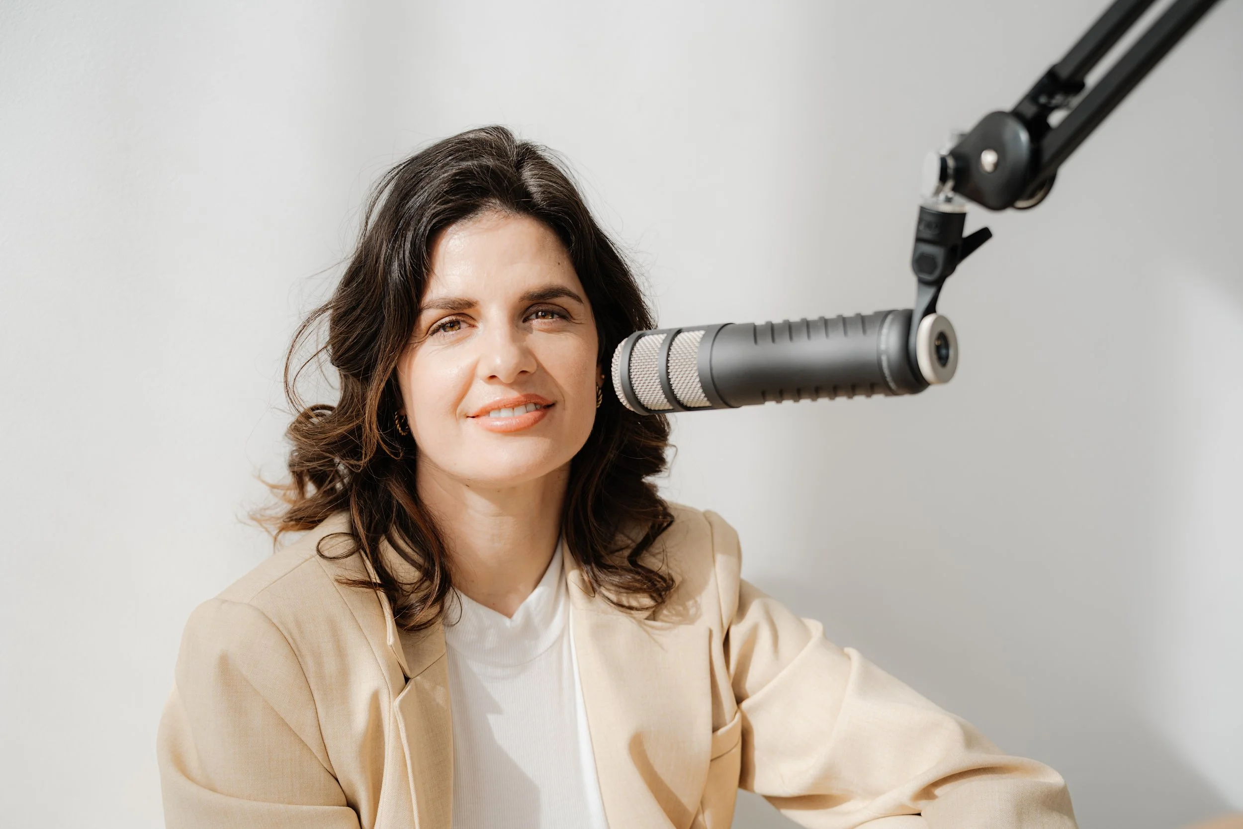 Eine Frau sitzt vor einem Mikrofon in einem Aufnahmestudio, trägt einen beigen Blazer und hat braune, gewellte Haare.