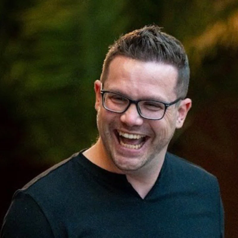 Ein lachender Mann mit Brille vor einem unscharfen grünen Hintergrund.