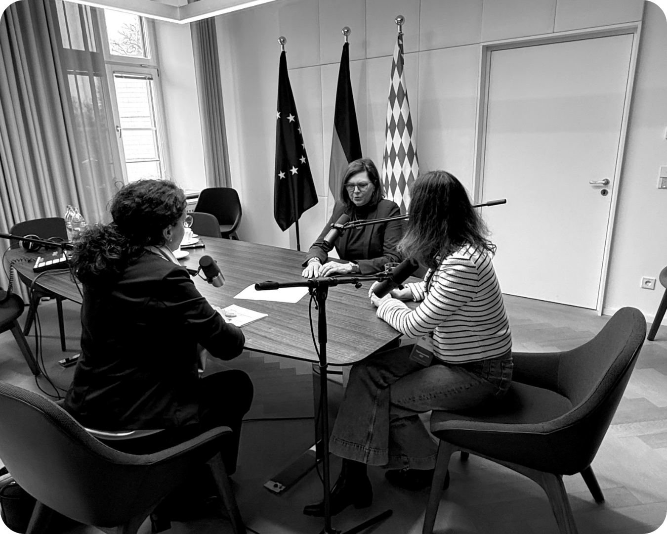 Drei Frauen sitzen an einem Tisch bei einer Aufnahme oder einem Interview, mit drei Mikrofonen vor ihnen, im Hintergrund stehen drei Flaggen, darunter die deutsche Flagge.