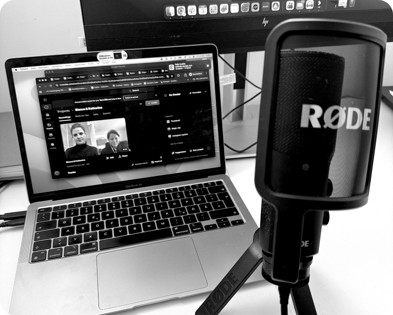Ein Laptop, ein Mikrofon (RØDE) und ein großer Bildschirm sind auf einem Schreibtisch zu sehen. Der Laptop zeigt eine Videokonferenz mit zwei Frauen auf dem Bildschirm.