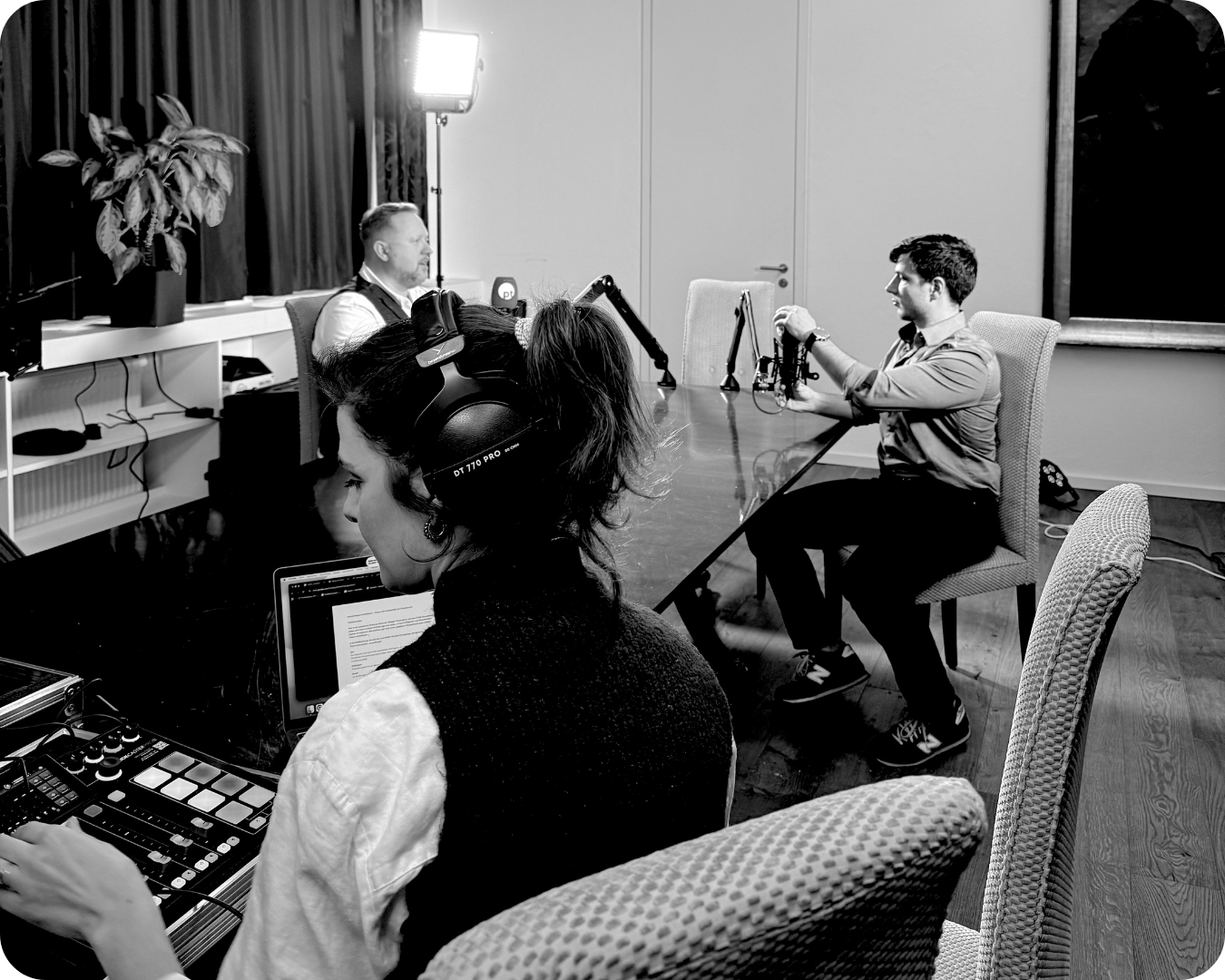 Drei Personen im Studio. Eine Person sitzt vor einem Laptop, trägt Kopfhörer, und arbeitet an einem Mixer. Zwei andere Personen sind im Hintergrund, eine sitzt mit einem Mikrofon und der andere mit einem Licht-Setup, vermutlich für eine Aufnahme.