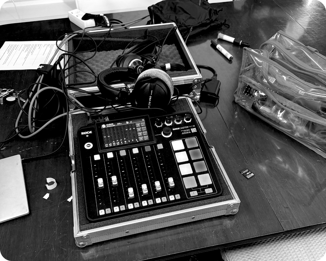 Tisch mit Audio-Equipment, darunter ein Mischpult, Kopfhörer, SD-Karten, Stifte, eine offene Tasche mit Zubehör und Kabel, alles in Schwarz-Weiß.