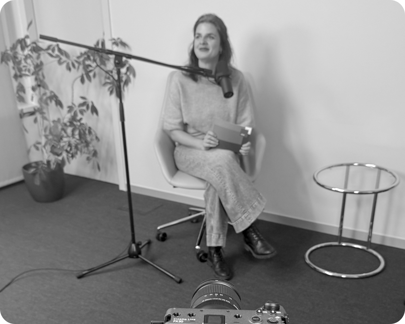Eine Frau sitzt auf einem Stuhl vor einer weißen Wand, hält ein Notizbuch und scheint eine Aufnahme zu machen, während sie vor einem Mikrofon sitzt, das an einem Mikrofonständer befestigt ist. Es gibt eine Pflanze in einem Topf im Hintergrund, eine k