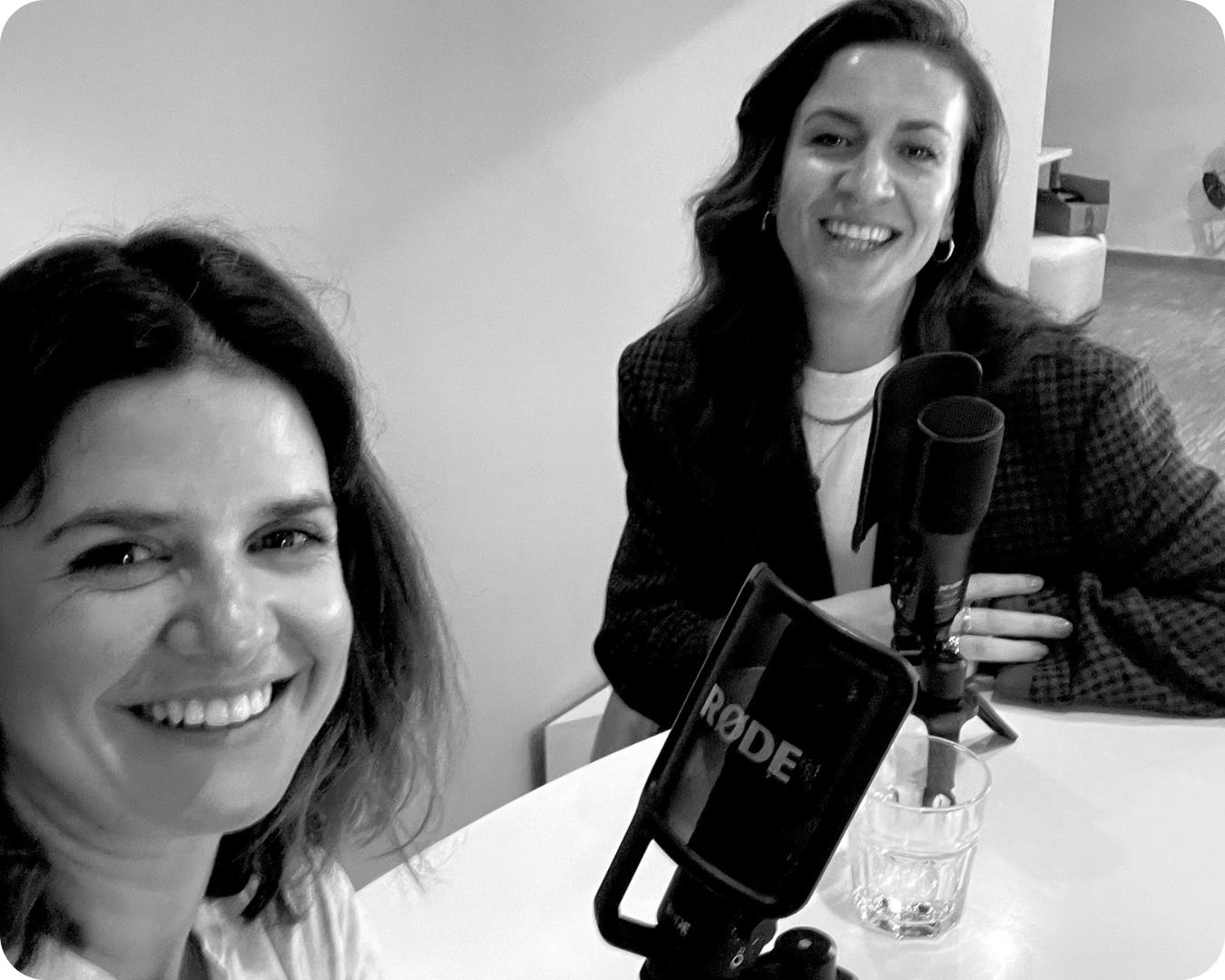 Zwei lachende Frauen vor einem Mikrofon in einem Raum, einer hat dunkle Haare, die andere helle Haare, es ist ein Schwarz-Weiß-Foto.
