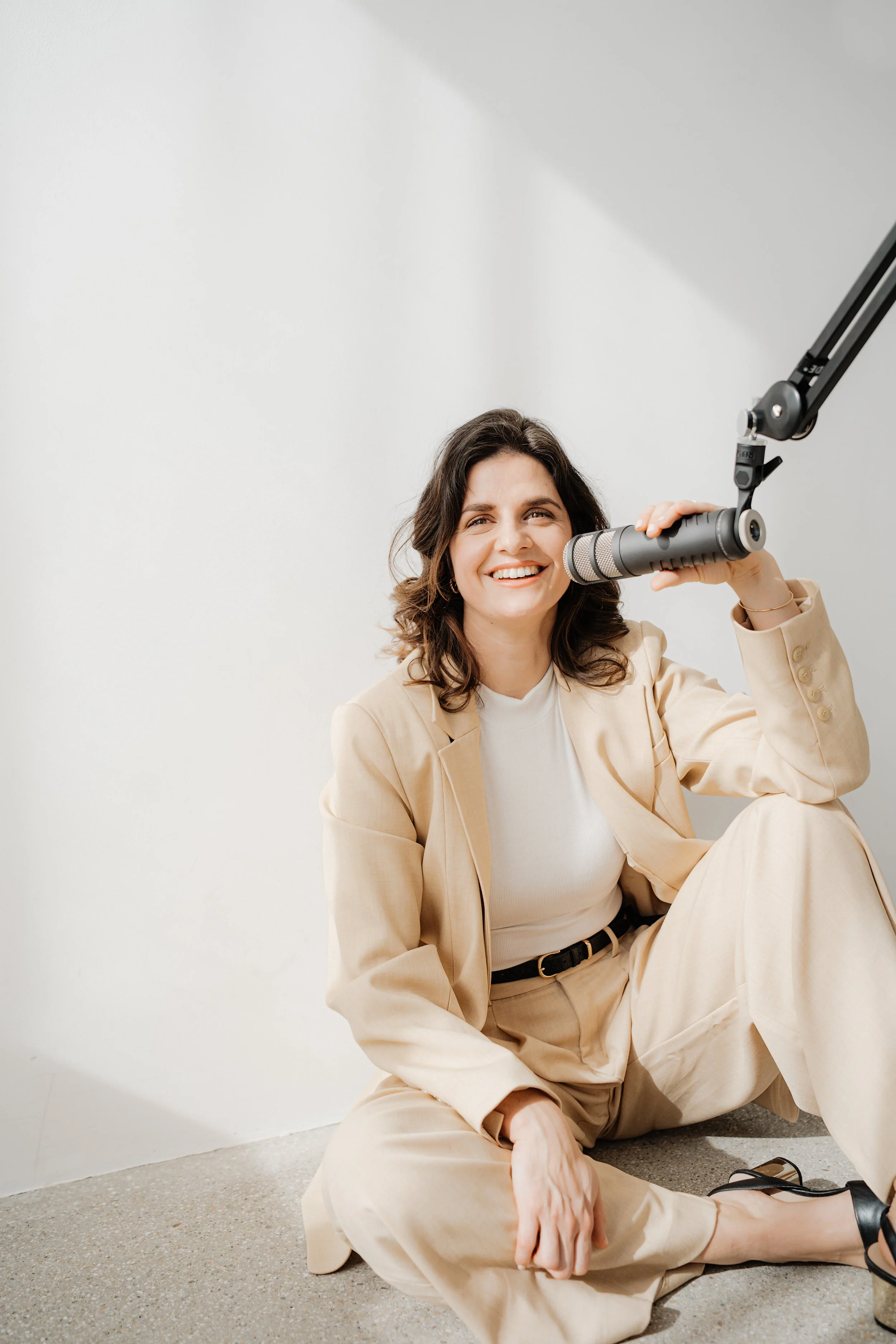 Eine lachende Frau in beige Anzug sitzt auf dem Boden, spricht in ein Mikrofon.