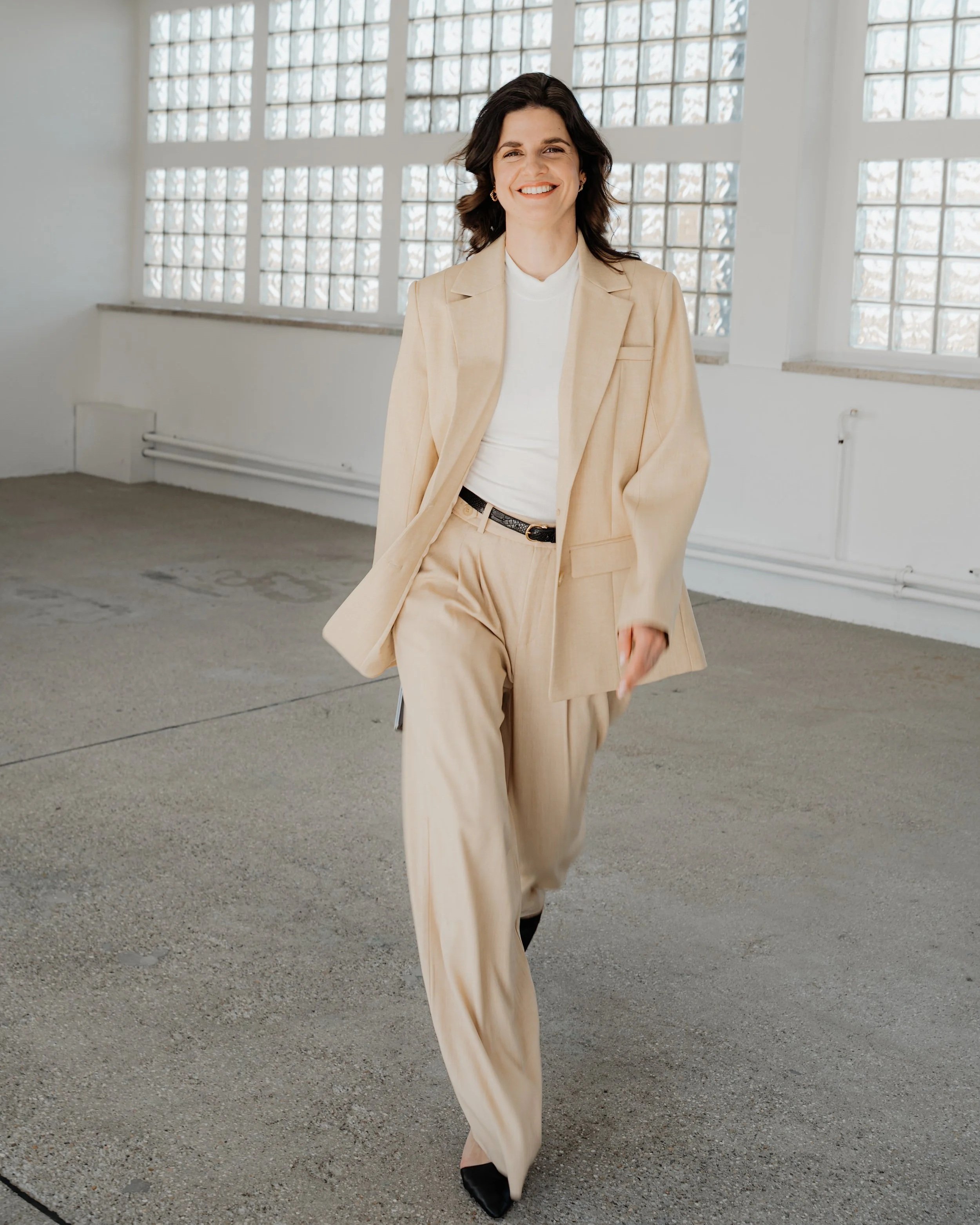 Eine lachende Frau in einem beige Anzug läuft in einem hellen, offenen Raum mit großen Glasfenstern.