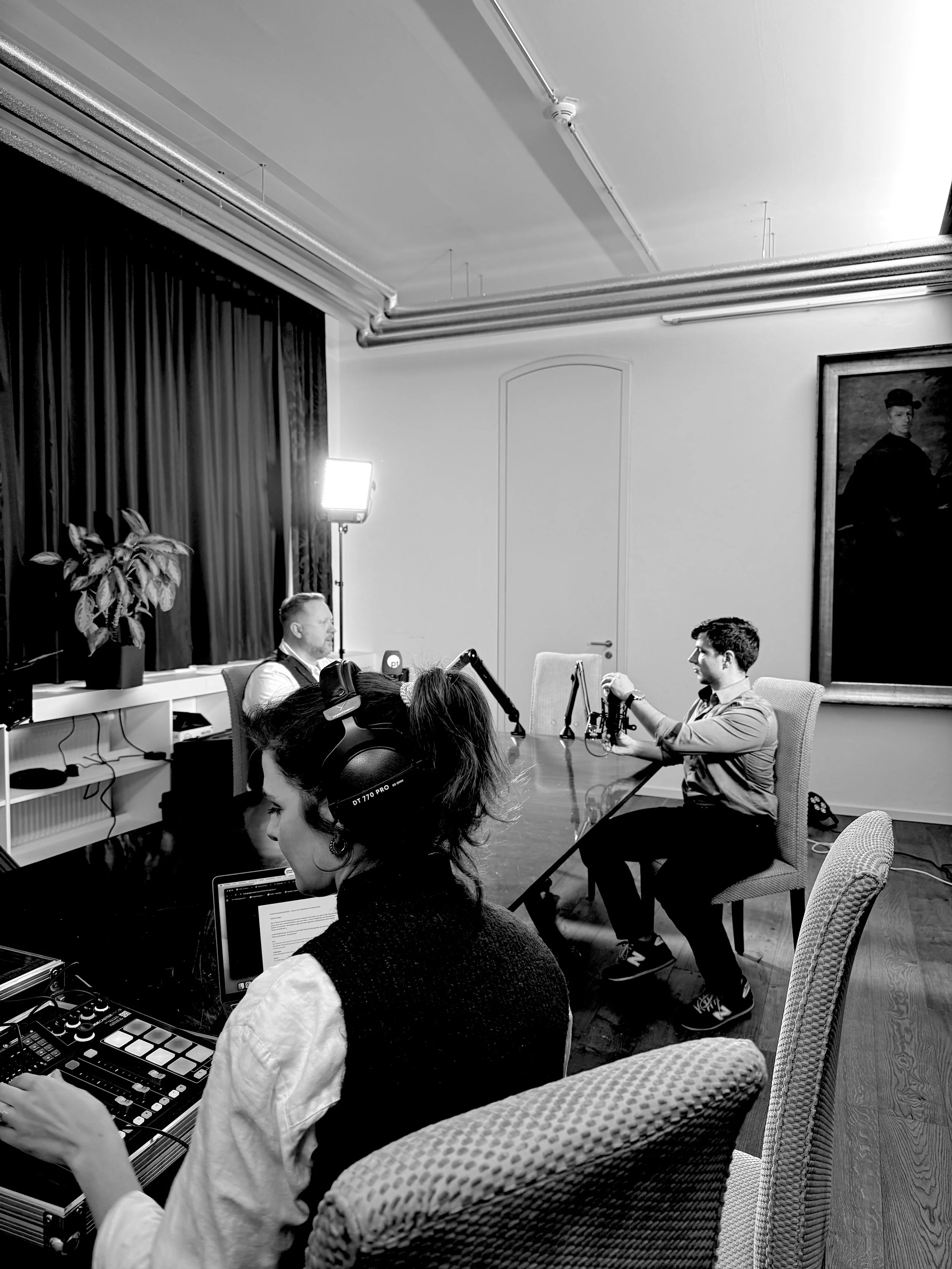 Drei Menschen sind in einem Raum bei einer Aufnahme oder Produktion. Der Mann vorne hält eine Kamera, die Frau im Vordergrund trägt Kopfhörer und arbeitet an einem Laptop, während der Mann im Hintergrund vor einem Mikrofon sitzt. Es gibt eine große Pflanze, eine große Wandmalerei und professionelle Beleuchtung.