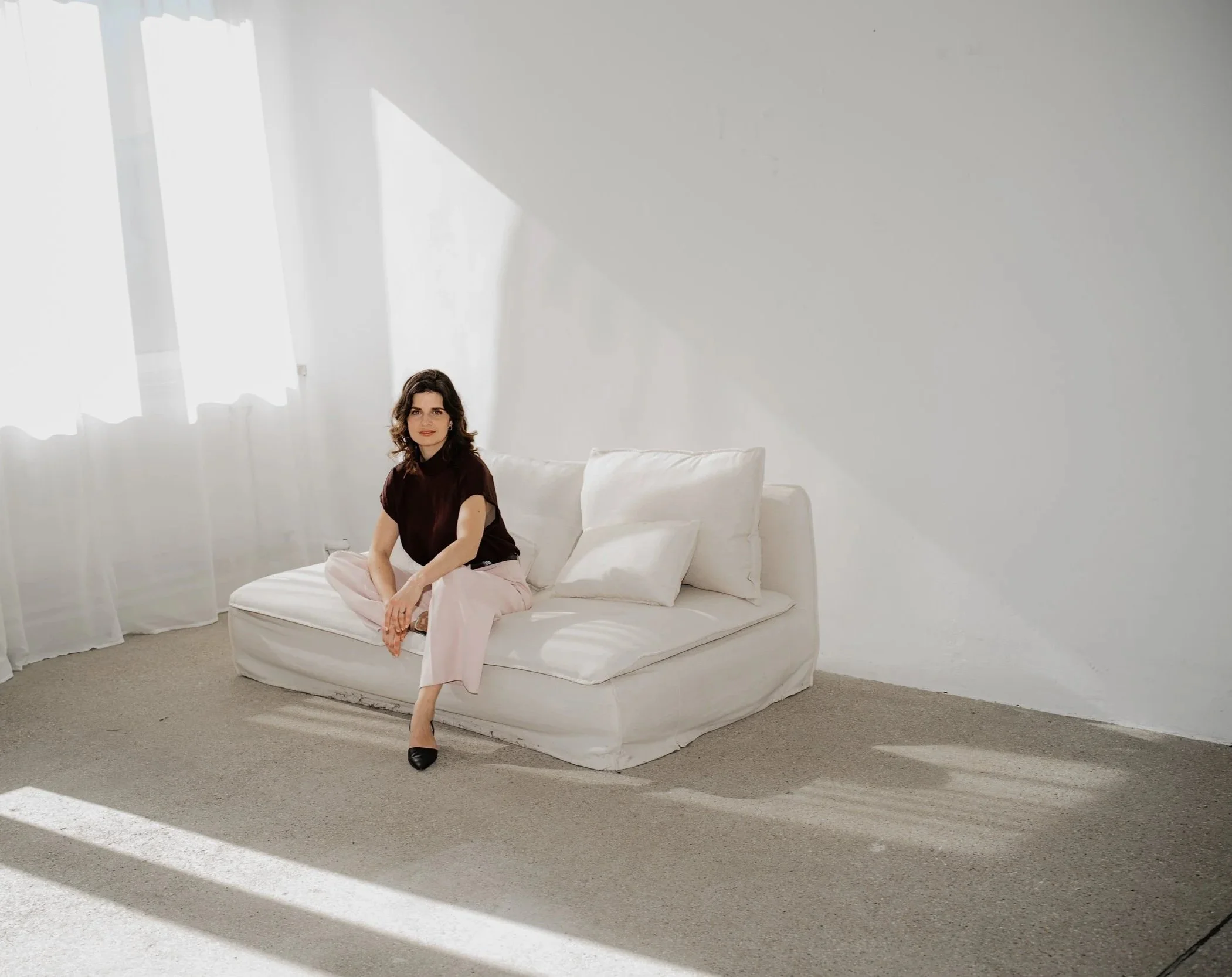 Eine Frau sitzt auf einem weißen Sofa in einem hell erleuchteten Raum mit weißen Vorhängen, die Sonnenlicht durchlassen.