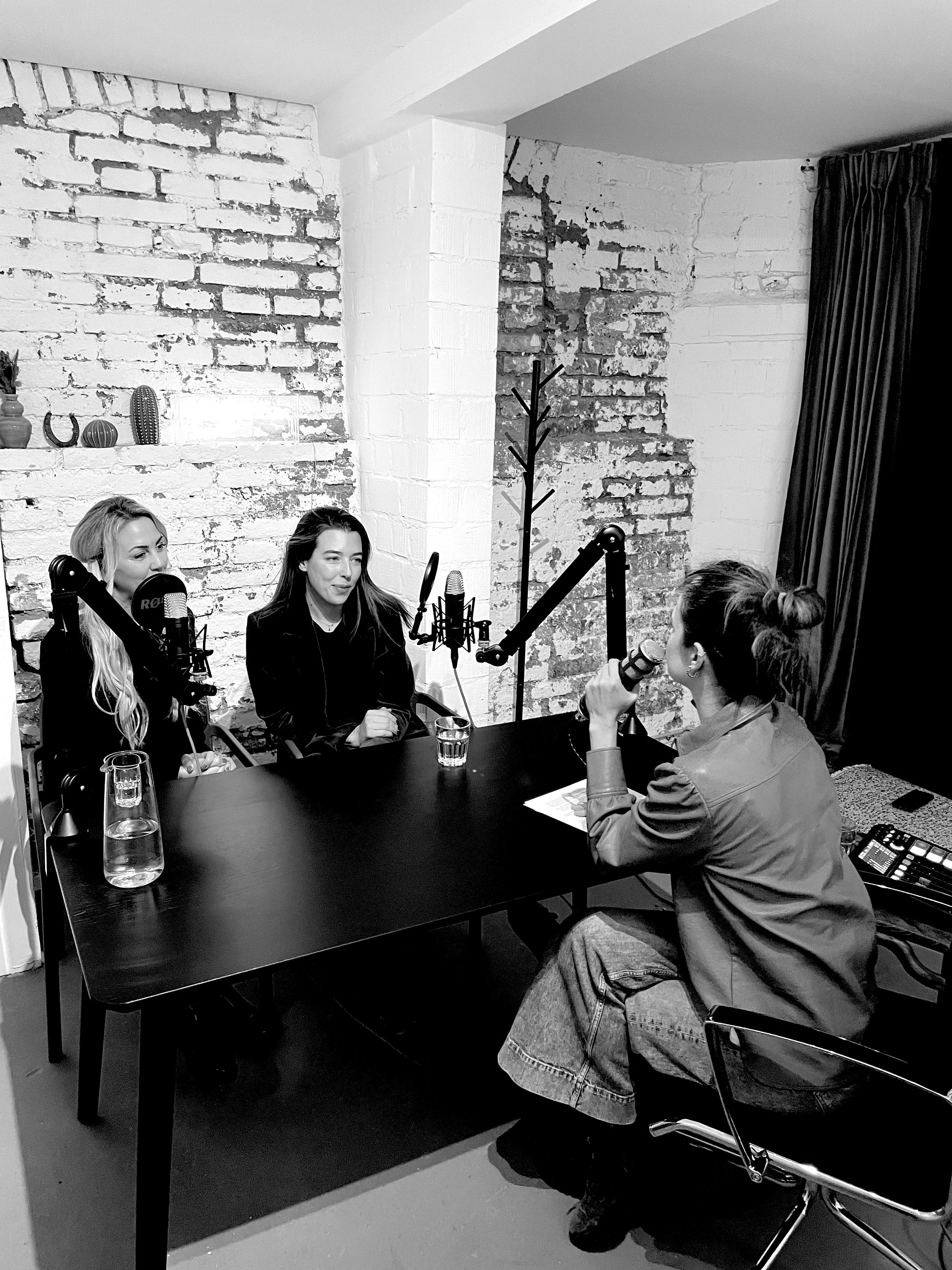 Drei Frauen sitzen an einem Tisch in einem Tonstudio, sprechen in Mikrofone und haben Wasserflaschen vor sich. Zwei Frauen sind zu sehen, eine spricht in ein Mikrofon, während eine andere zuhört.