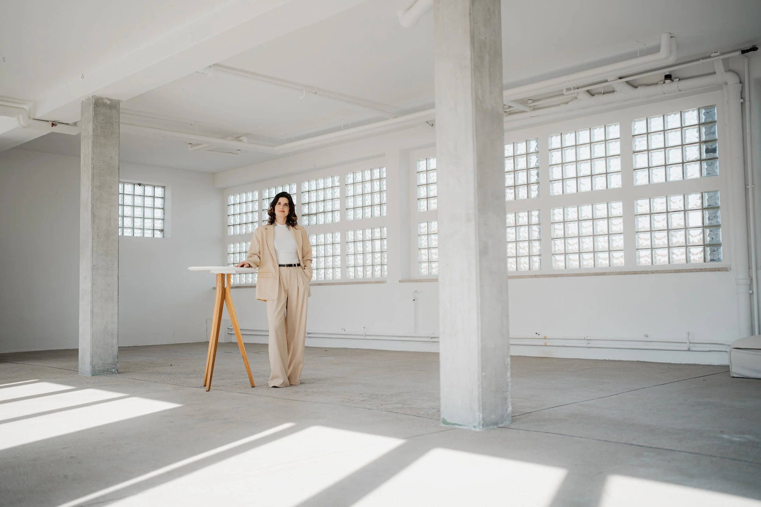Frau in beige Anzug steht in einem hellen, leerem Raum mit großen Glasblockfenstern, die Sonnenlicht hereinlassen.