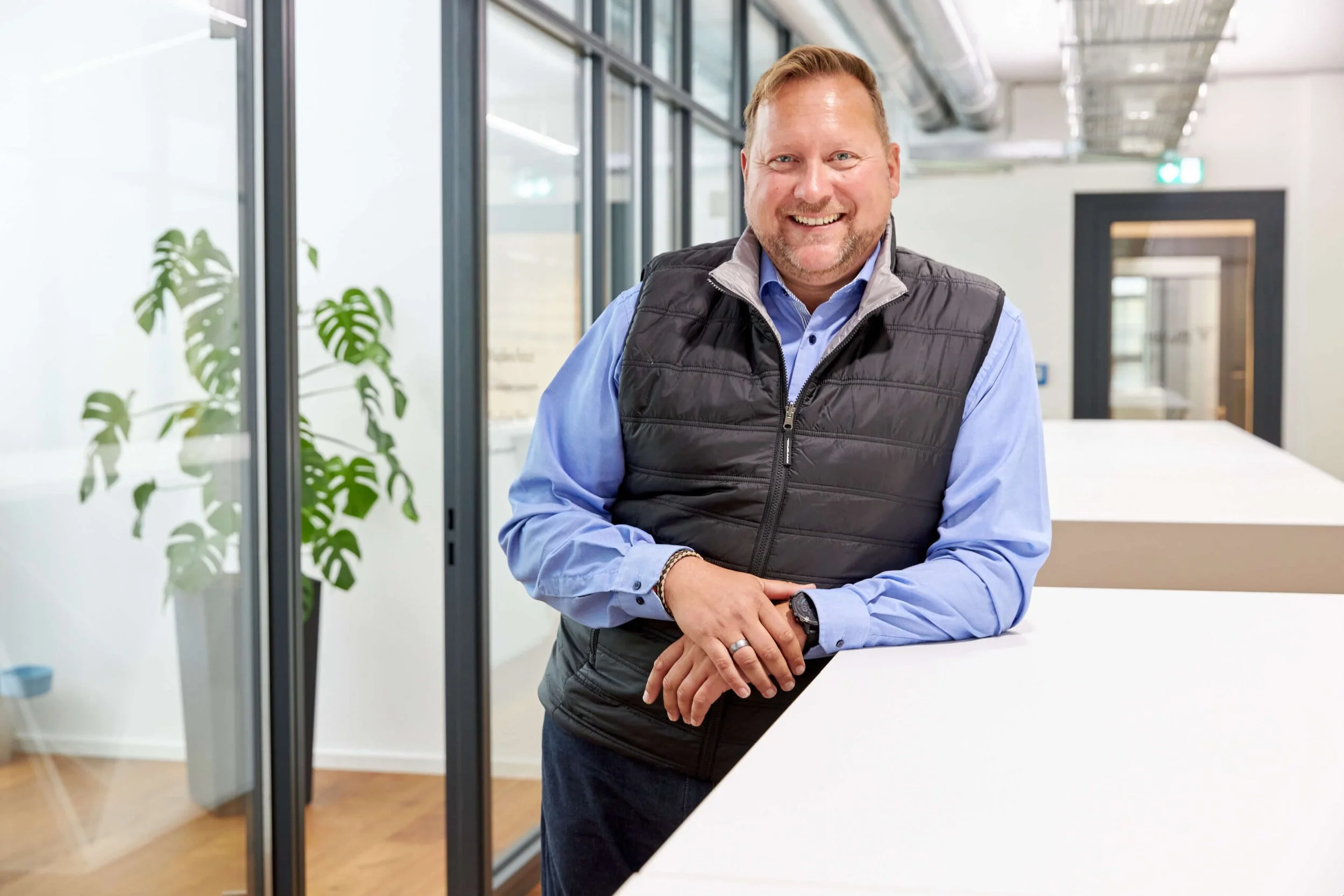 Lächelnder Mann in einem modernen Büro, trägt eine blaue Hemd und eine schwarze Weste, steht an einem weißen Tresen.