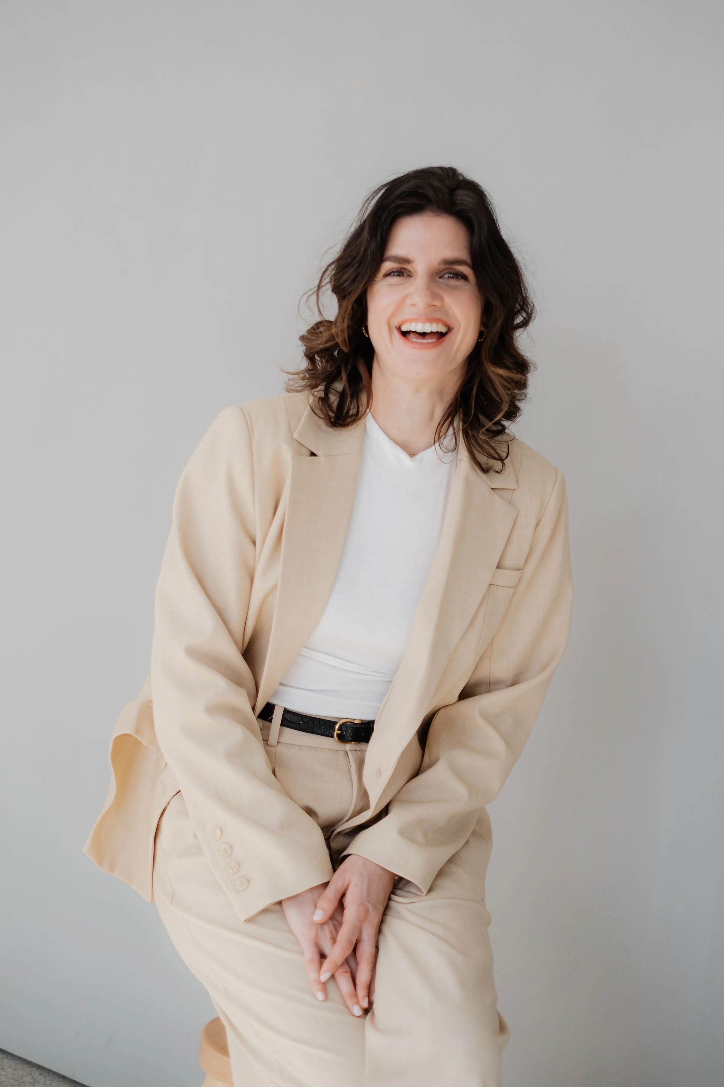 Junge Frau mit braunen, lockigen Haaren, lachend, in beige Anzug und weißem Shirt, vor hellem grauem Hintergrund.