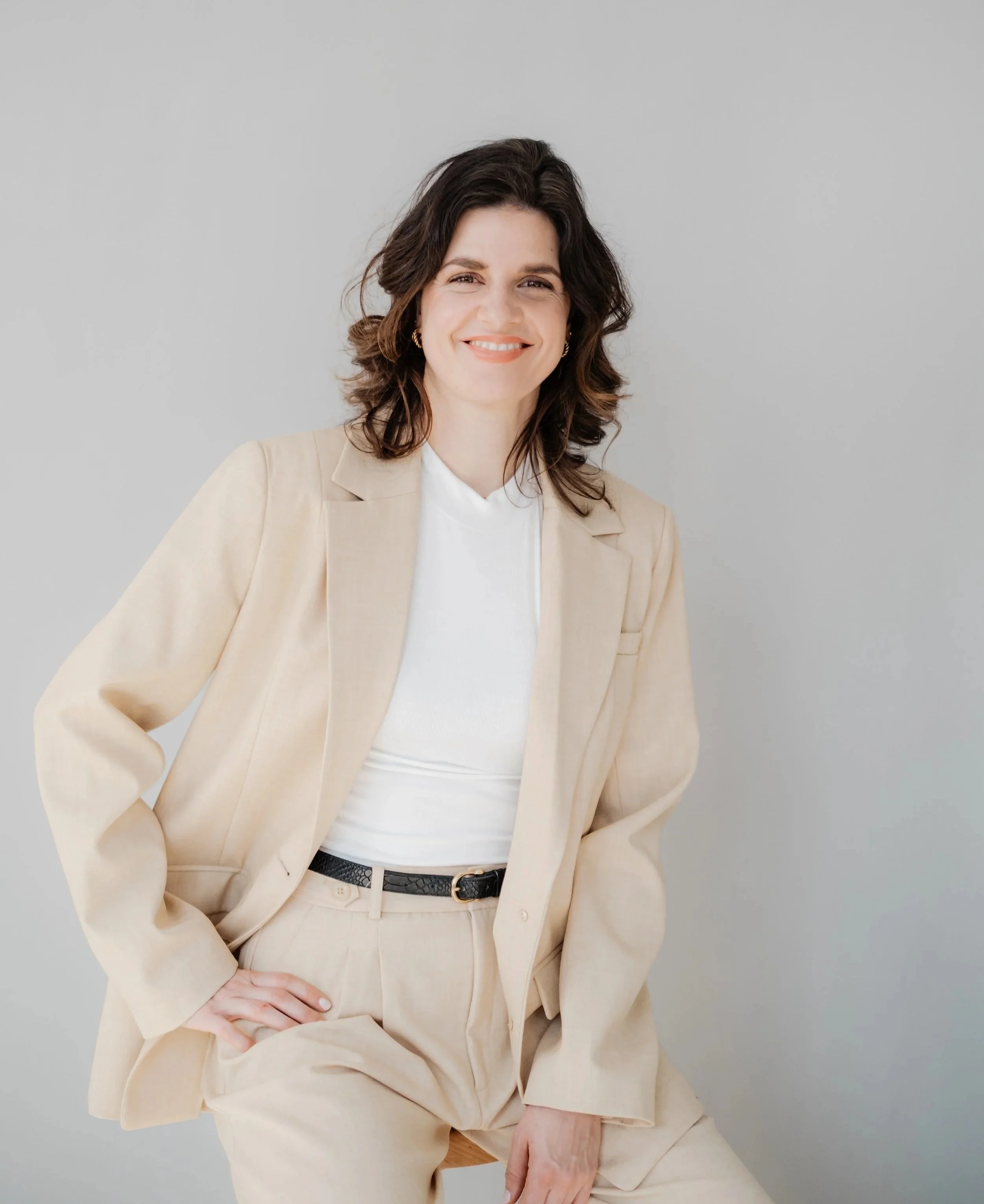 Frau in beige Anzug mit weißem Hemd sitzt vor grauem Hintergrund, lächelt, mit Hand auf der Hüfte.