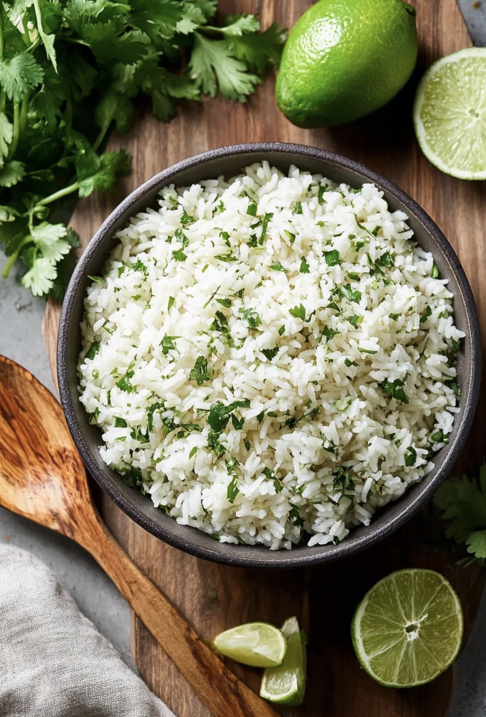 A bowl of cooked white rice garnished with chopped cilantro, surrounded by fresh cilantro, lime, and lime wedges on a wooden surface.