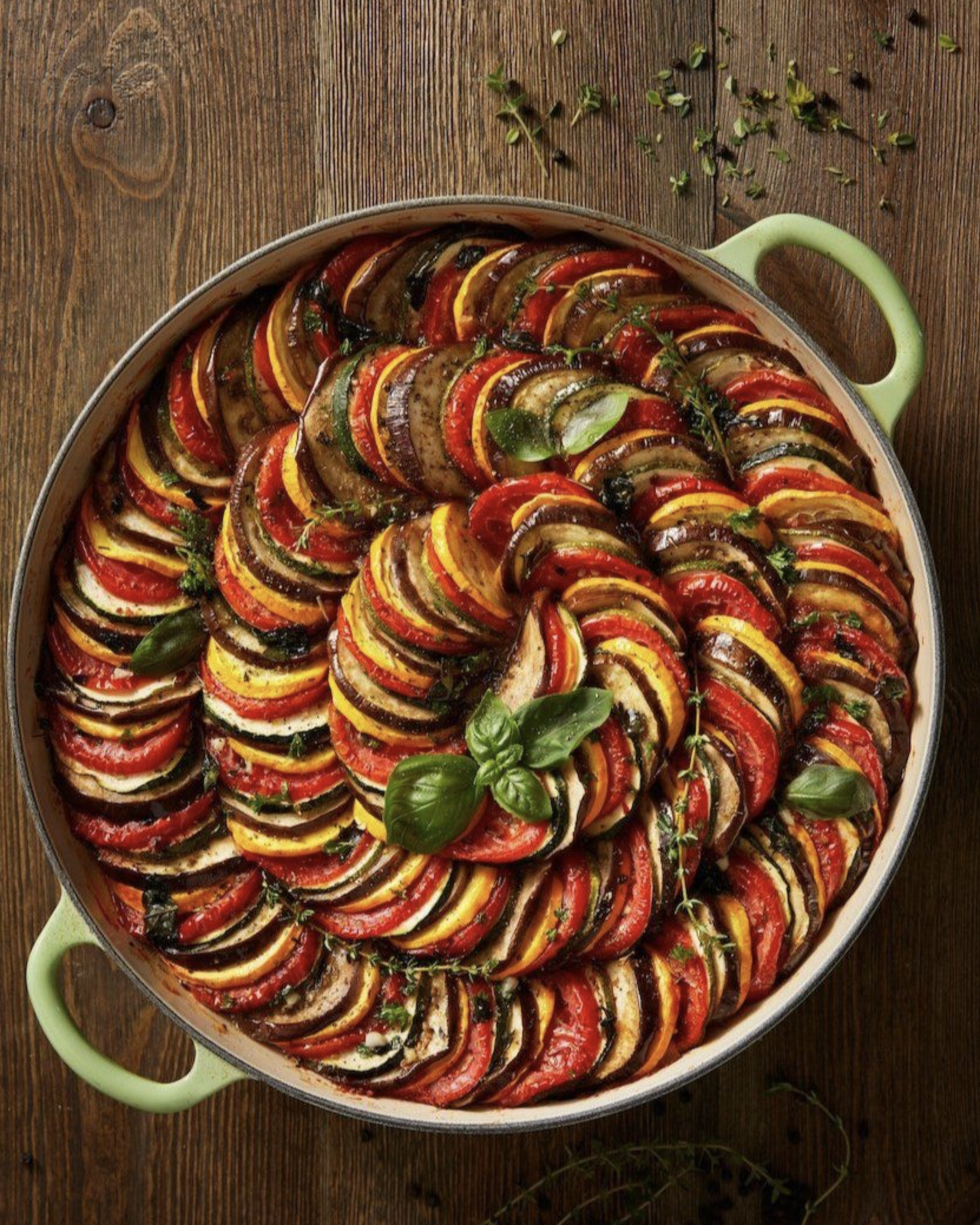 A round baking dish filled with a spiral of sliced, roasted vegetables including tomatoes, zucchini, eggplant, and bell peppers, garnished with fresh basil and thyme, on a wooden surface.