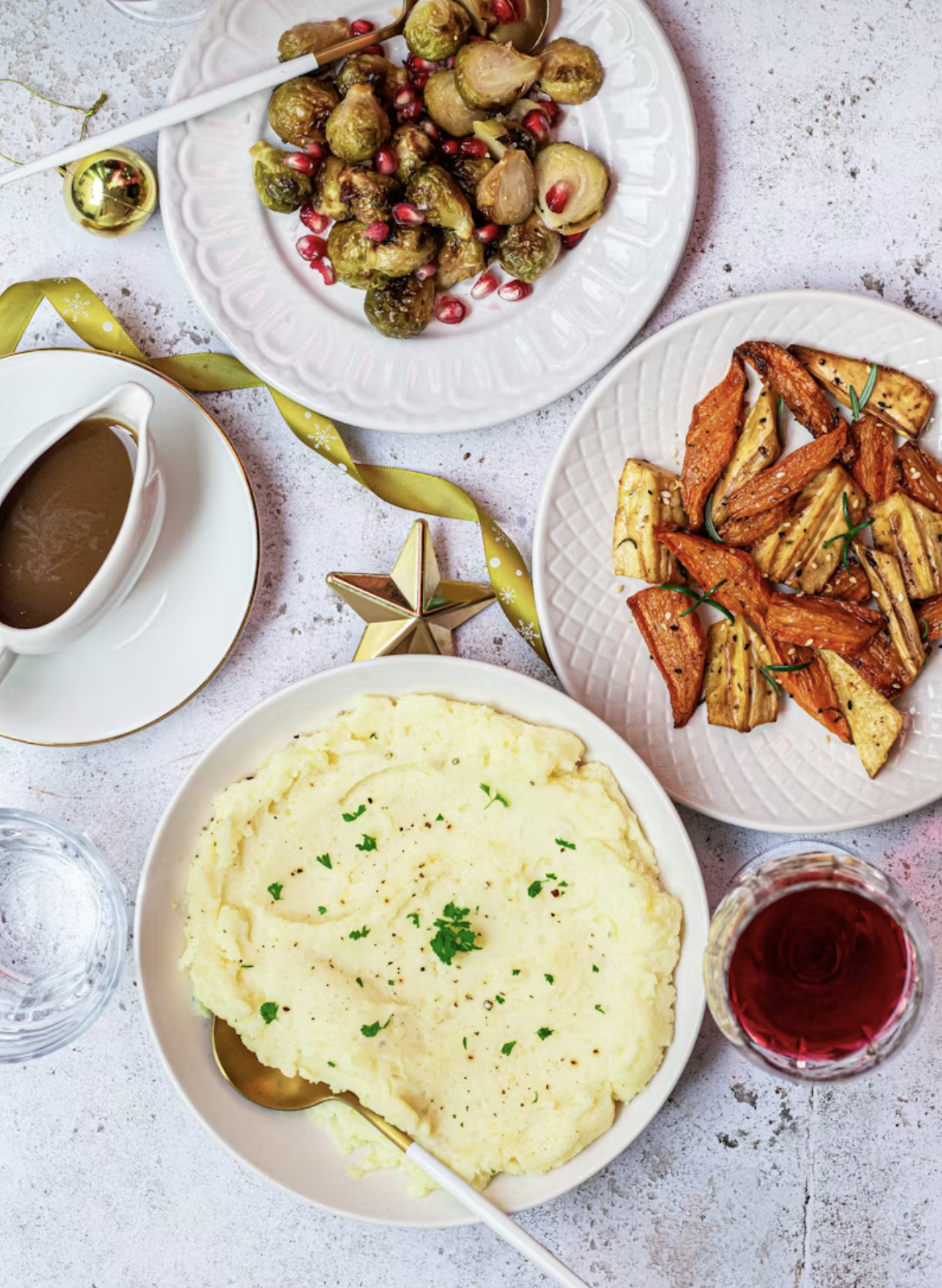 Holiday meal with mashed potatoes, roasted vegetables, Brussels sprouts with pomegranate seeds, a glass of red wine, a cup of coffee, and a small glass of water on a festive table.