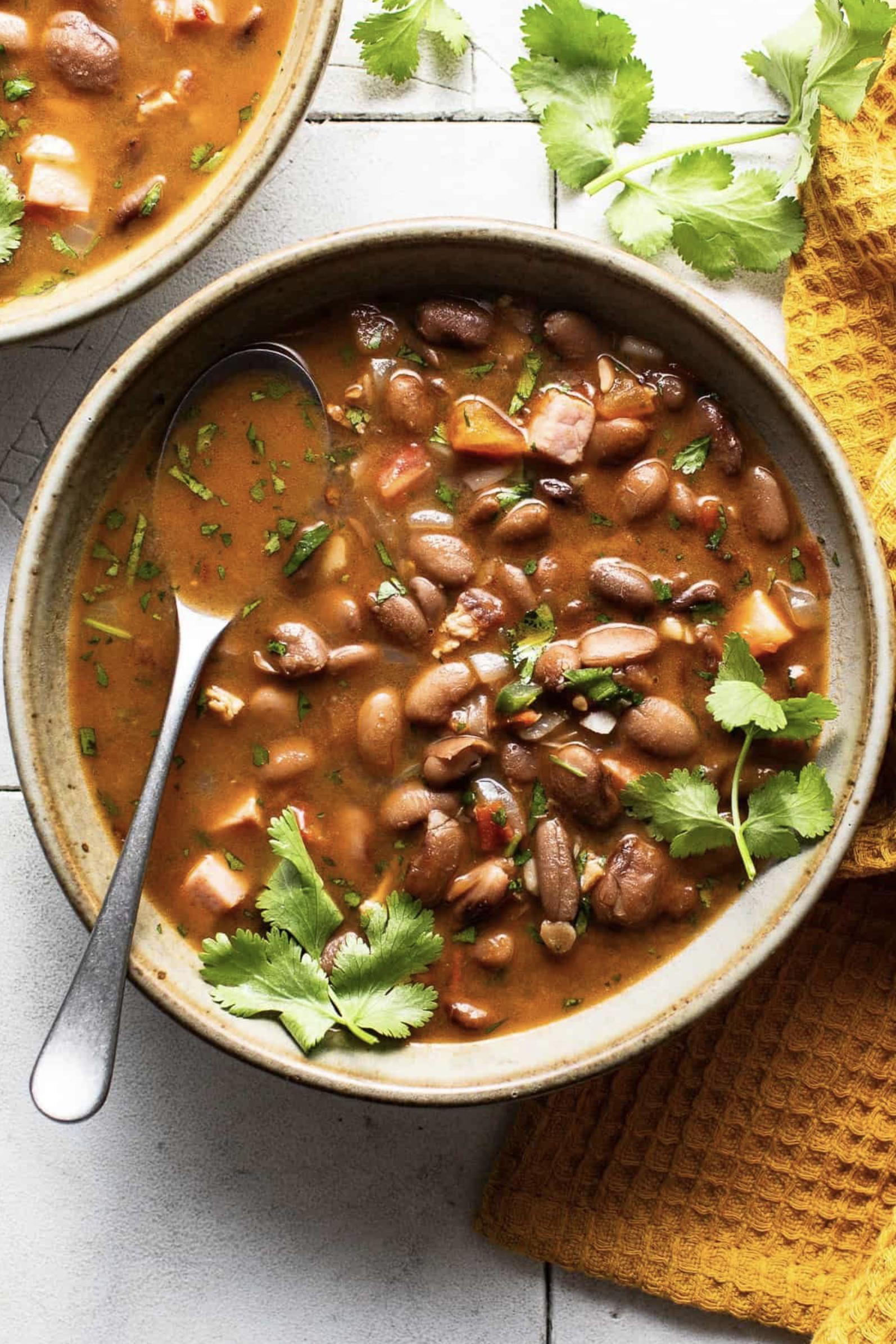 Bowl of black bean soup garnished with cilantro leaves, with a metal spoon, on a white surface with a mustard yellow cloth nearby.