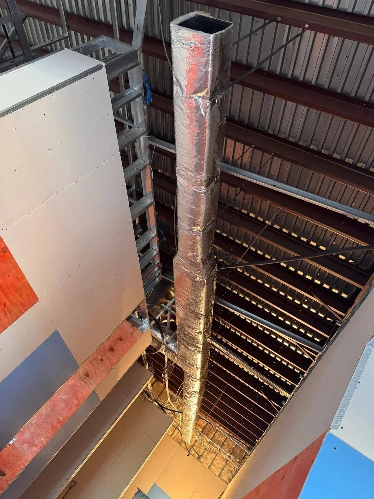 A construction site ceiling showing metal beams, a large insulated duct, and an unfinished wall with drywall panels.