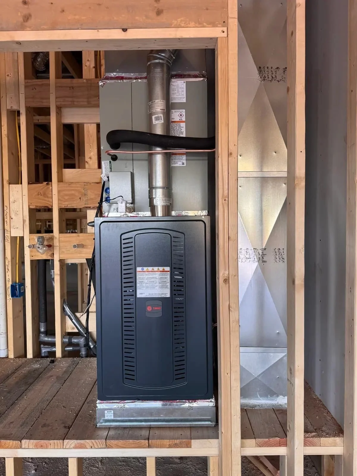 A furnace and air handler unit installed in a building under construction, with exposed wooden framing and an unfinished floor.