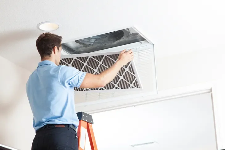 A technician in a blue shirt is repairing an HVAC air conditioning unit mounted on a ceiling, standing on a ladder in a room.