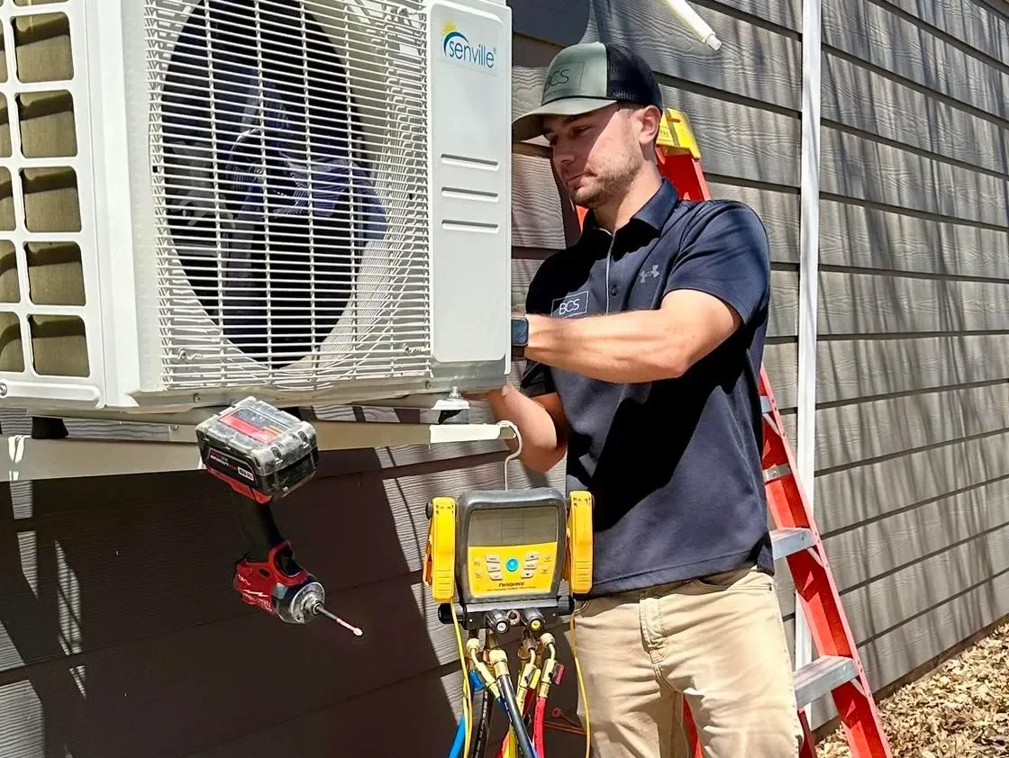 A technician installing or servicing an outdoor air conditioning unit on the side of a building, with a ladder and HVAC tools nearby.