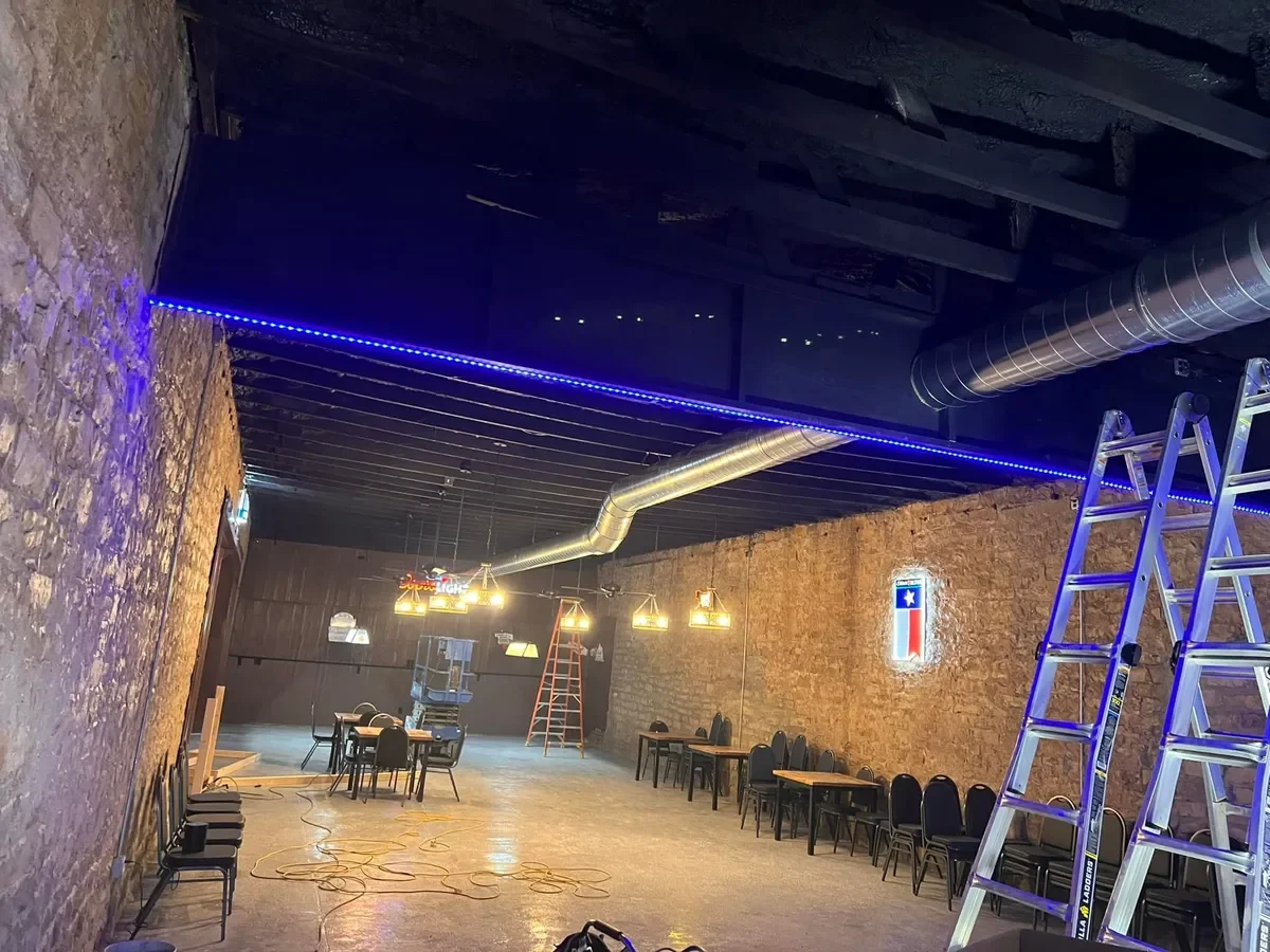 Interior of a commercial space under renovation with exposed brick walls, a ladder, chairs, tables, hanging pendant lights, a LED strip light along the ceiling, and scaffolding.