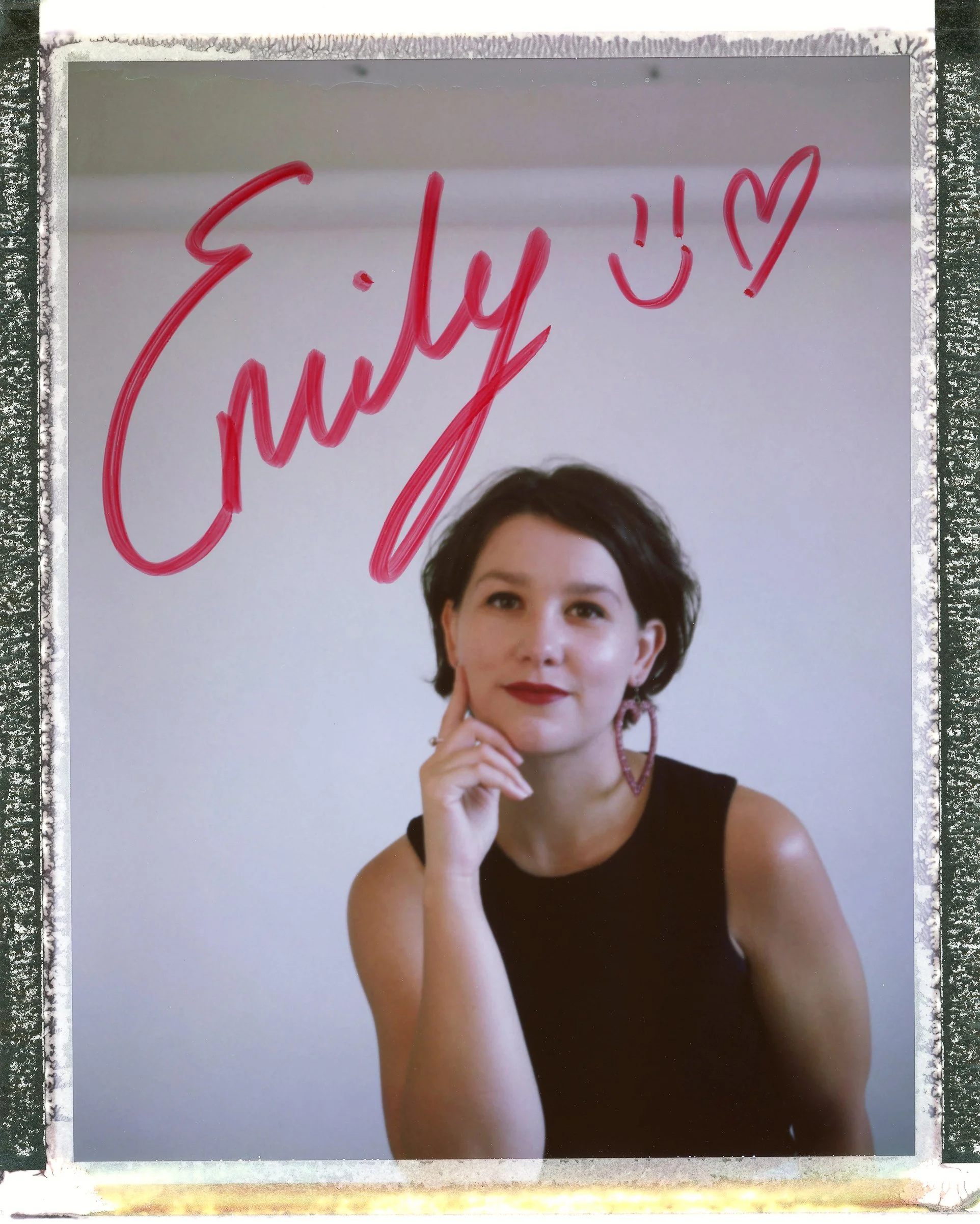 A woman with dark hair, wearing a black sleeveless top and pink earrings, is posing with her chin resting on her hand, looking at the camera. Above her, the phrase 'July ' and a smiley face and heart are written in pink marker on a reflective surface.