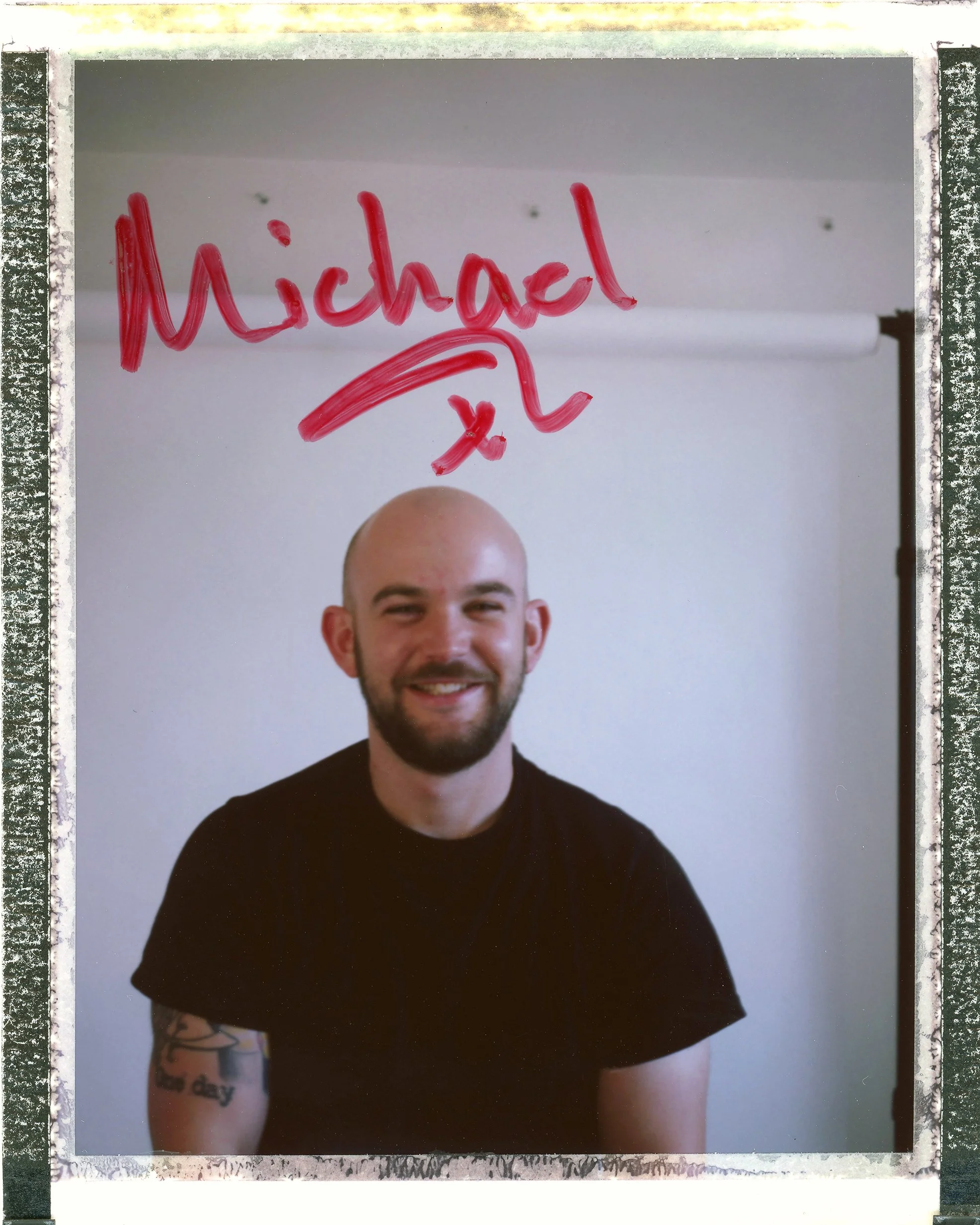 A smiling man with a beard and tattoos on his left arm, sitting in front of a whiteboard with the name 'Michael' written on it in red marker.