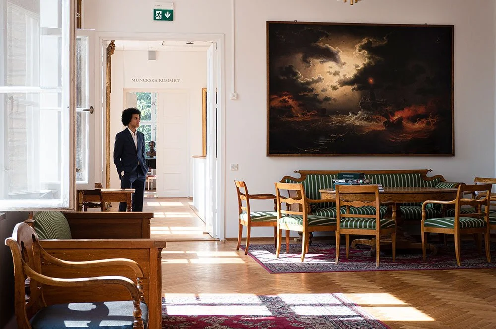 Ett rum med ett möblerat sällskapsområde och en man i kostym som står i dörröppningen, konstnärlig målning på väggen, samt stora fönster som släpper in ljus.