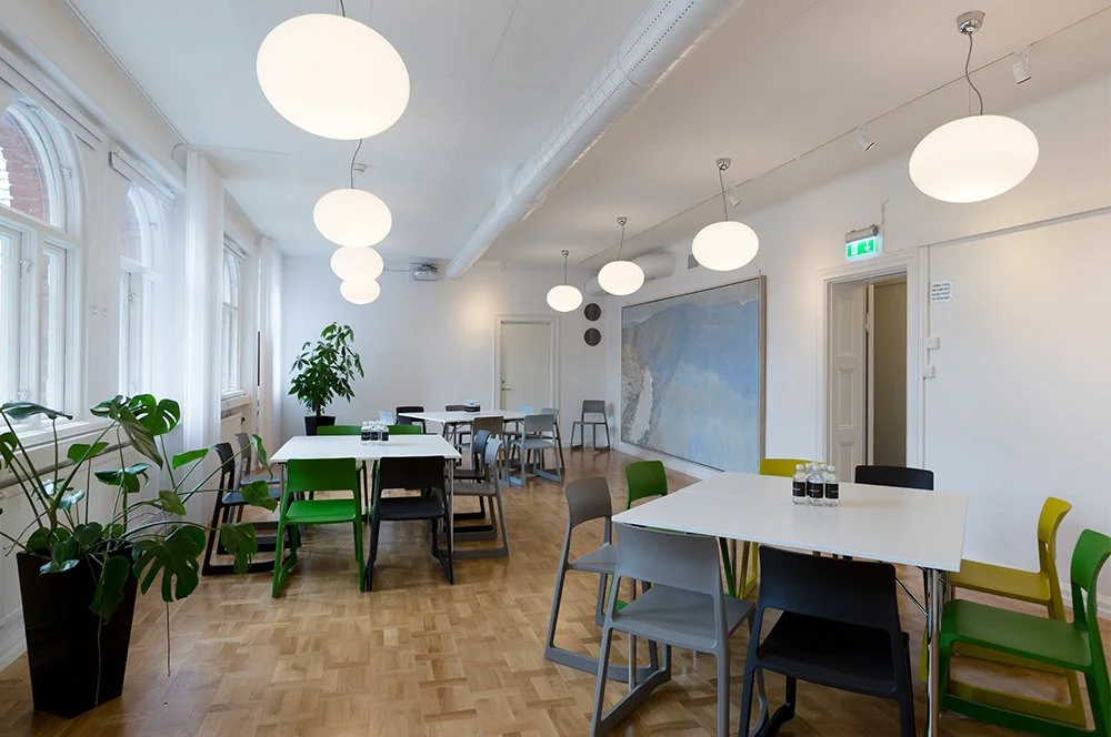 Ett rent och ljust konferensrum eller mötesrum med vita väggar, stora fönster och golv av ljus trä, inrett med flera bord och stolar i olika färger samt gröna växter.