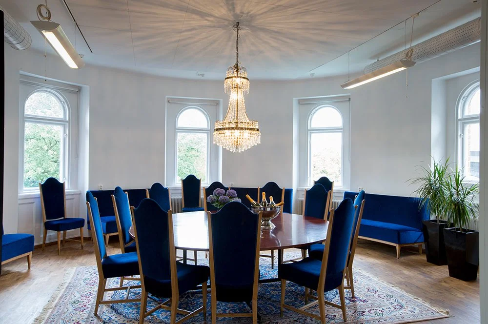 Ett rundbord med åtta blå klädda stolar i ett elegant mötesrum, med ett kristallkronljus i taket, stora fönster och ett växt på sidan.