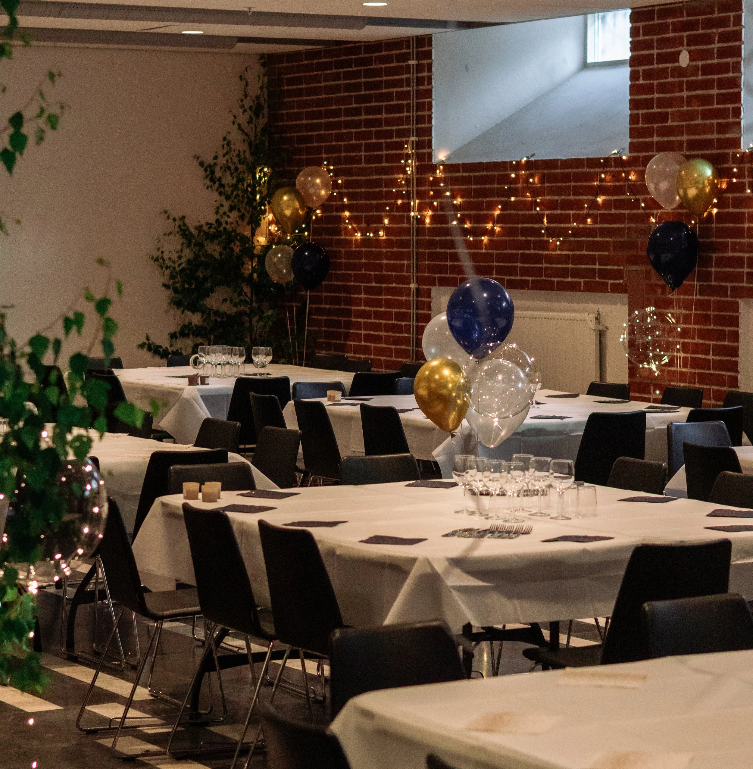 Källarsalen festpyntat i ett rum med runda bord, svarta stolar, ballonger i guld, svart, blå och vit, dekorativa ljusslingor och ett julgran i hörnet, fönster och en tegelvägg i bakgrunden.
